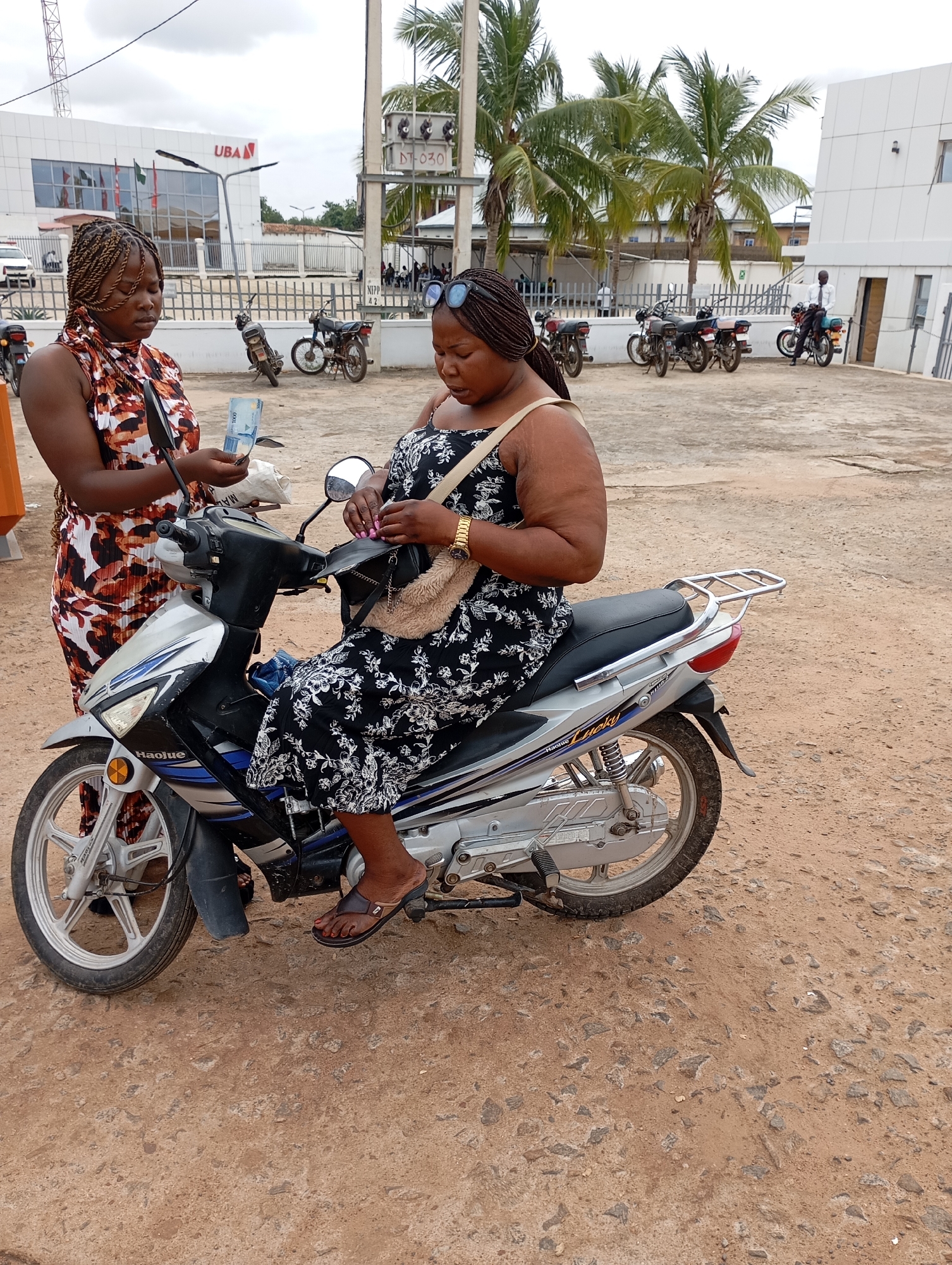 A Nigerian woman, wearing a black and white floral dress, sits on a silver motorcycle in a dirt lot. She is holding a beige purse and has her hands near the handlebars. Another woman, wearing a patterned sleeveless dress, stands to the left of the motorcycle, holding money and a white pouch. In the background, a UBA bank building is visible, along with several parked motorcycles and palm trees. The scene appears to be outside a commercial area in Shendam, Nigeria.