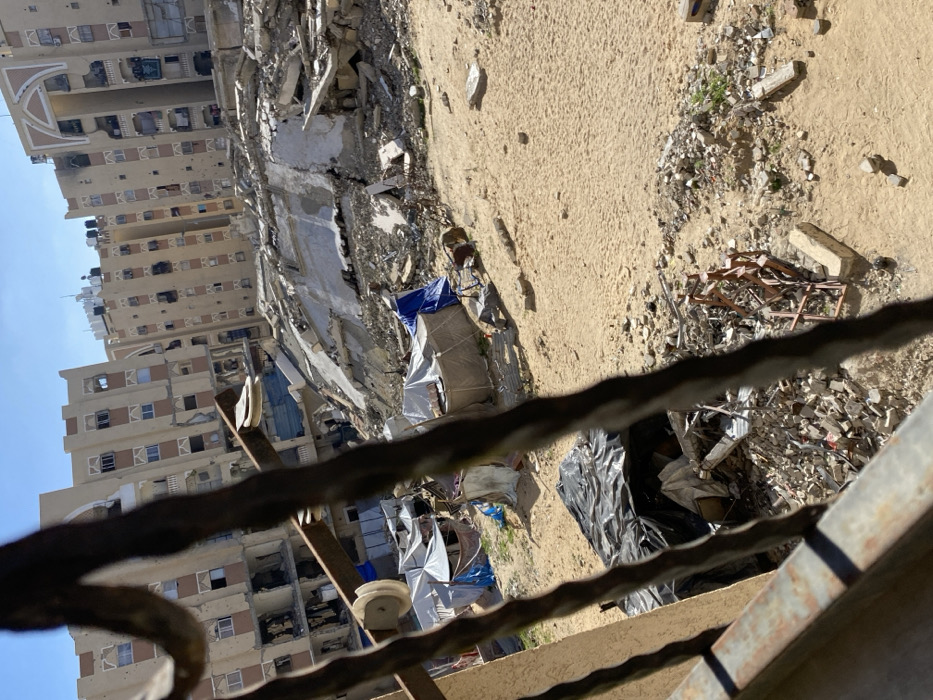 The image depicts a devastated urban landscape in Khan Yunis, Palestinian Territory, captured from an elevated perspective, likely through a metal window grille or balcony railing in the foreground. The scene is dominated by extensive structural damage and debris under a clear blue sky.
In the immediate foreground, dark, twisted metal bars, possibly part of a protective grille, partially obscure the view. Below these bars, a dark tarp covers a pile of rubble and debris on sandy ground.
The midground features an open expanse of sandy, light brown earth, interspersed with piles of concrete fragments, twisted rebar, and assorted wreckage. On this ground, a cluster of makeshift shelters, predominantly constructed from light-colored tarpaulins, some with blue tarp accents, are visible. One shelter appears somewhat conical or dome-shaped. Near these shelters, a light brown dog is seen lying on the sand.
Further back, a massive mound of collapsed building material, consisting of concrete slabs, rebar, and other structural elements, forms a significant barrier across the scene. Behind this immense rubble pile, several multi-story residential buildings stand, exhibiting severe damage. These beige-toned buildings feature numerous shattered windows, missing wall sections, and visible impact points or scorch marks on their facades. Black water tanks are visible on the rooftops of these damaged buildings. One building's upper section includes a distinctive arched architectural detail.