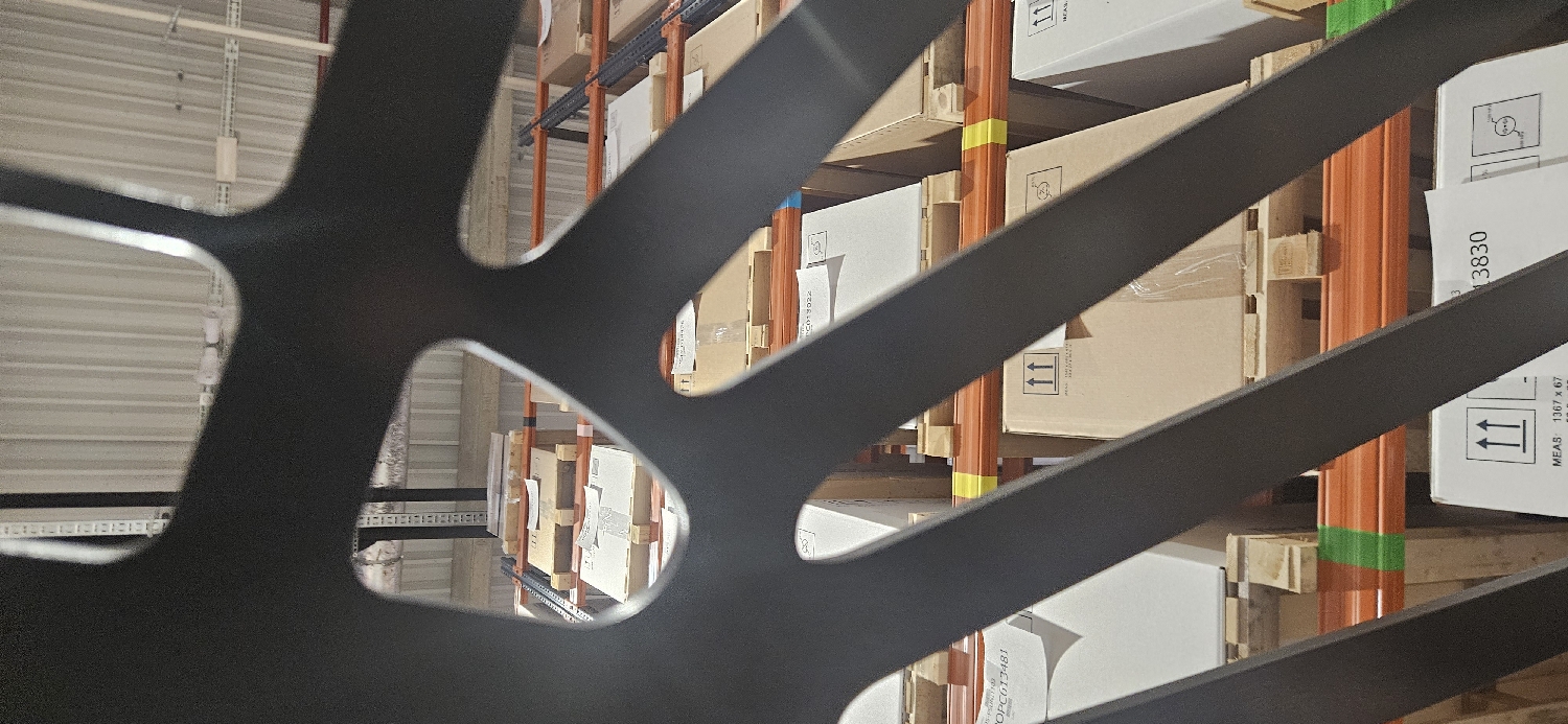 The image depicts an indoor scene, likely a warehouse or large storage facility, viewed through a dark, irregularly shaped object in the immediate foreground. This object, possibly made of metal, has multiple curved arms or spokes that partially obscure the background, creating an abstract, framed view of the items behind it.
The primary subjects in the background are rows of industrial racking systems, stacked high with numerous cardboard and white boxes. The racking features prominent orange vertical support beams, some of which have small strips of yellow or green tape wrapped around them. Many boxes are resting on wooden pallets. The overall lighting suggests a well-lit interior, either from overhead fixtures or natural light filtering in. The far background shows a corrugated metal ceiling or wall structure.
No human activity is visible, presenting a static snapshot of a storage environment. The image has a slight tilt, making the vertical elements of the racking appear slightly diagonal.
Notable details include:
* **Visible Text:** On several white boxes, upward-pointing double arrows are clearly visible, indicating handling instructions. Other text fragments include "3830", "1367 x 67", and "MEAS". Pieces of white paper attached to some boxes show alphanumeric codes such as "OPC13022" and "OPCG13481".
* **Colors:** The palette is dominated by dark grey/black in the foreground, with browns, whites, and oranges in the background. Light grey tones are visible on the ceiling/walls.
* **Location Cues:** The racking systems, stacked boxes, and industrial ceiling strongly suggest a commercial or industrial storage facility, such as a warehouse. The location is an indoor facility in Plovdiv, Bulgaria.