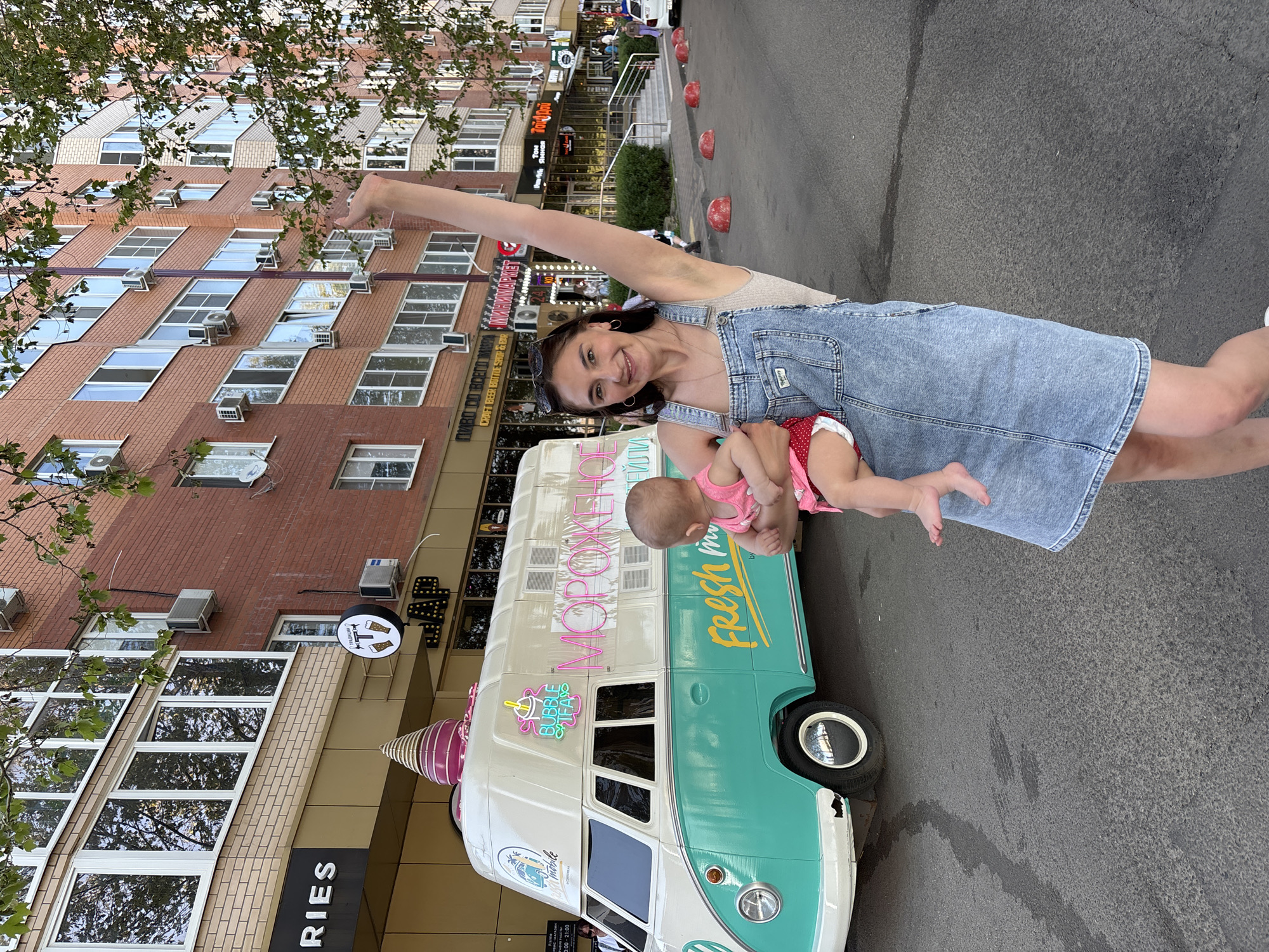A woman holds a baby next to a vehicle in an urban environment. The woman has dark hair pulled back and is wearing a denim dress. She has one arm raised. The baby is wearing a pink outfit. The vehicle is decorated in pastel colors with images of ice cream and the words "Fresh" and "Bubble Teas." Behind the vehicle, there is a brick building with many windows and air conditioning units. There are signs on the building for "CRAFT BEER BUTTLE SHOP & BAR" and "МИНИМАРКЕТ." The street appears to be in Krasnodar, Russia, as indicated by the Russian writing on the vehicle and signs. The street is paved, and there are some red bollards along the curb.