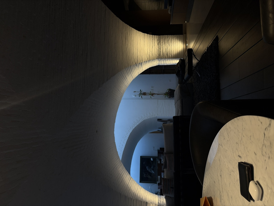 The image depicts a unique interior featuring a series of arched, white brick passageways. The arches lead to rooms with soft lighting, displaying furniture like sofas and a framed picture on the wall. The visible part of a room on the right includes a table with a wallet, computer mouse, and chair. The perspective creates a tunnel vision effect, emphasizing the architectural design and the depth of the space.