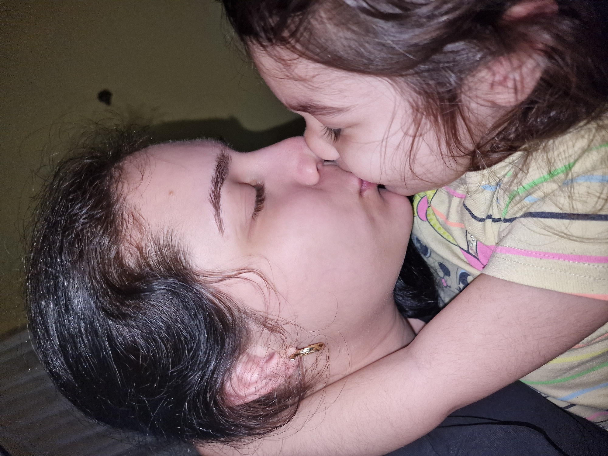 The image presents a close-up, intimate portrait of two individuals, likely an adult and a young child, sharing a tender kiss on the lips. The scene appears to be indoors, against a plain, dark olive-green background.
The adult, on the left side of the frame, has dark, slightly wavy hair and fair skin. Their eyes are closed, conveying a sense of warmth and focused affection. A small, golden hoop earring is visible in their left ear. They are wearing a dark-colored garment.
The child, positioned on the right, leans in to kiss the adult. The child also has dark, flowing hair and fair skin, and their arm is wrapped around the adult's neck or shoulder in a hug. They are wearing a light-colored t-shirt adorned with colorful horizontal stripes in shades of yellow, blue, green, and pink. A partial graphic resembling cartoon characters, possibly Mickey Mouse ears, is visible on the pink stripe of the shirt.
The overall atmosphere of the image is one of profound love, closeness, and tenderness. The lighting is soft and subdued, typical of an indoor setting, but does not provide specific clues about the time of day. There are no visible location cues within the image to suggest Byala Slatina, Bulgaria. No text is visible in the media file.