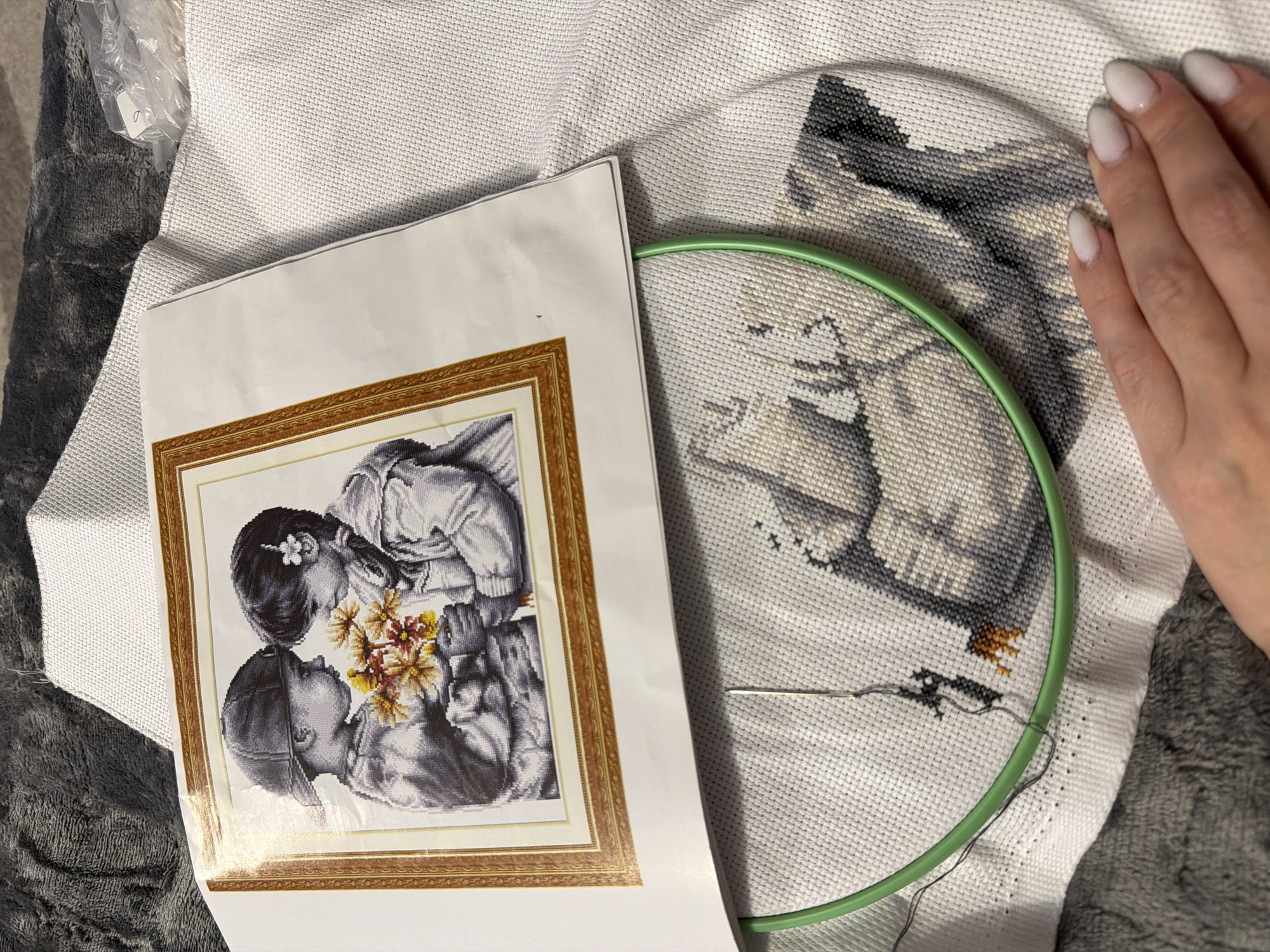The image shows a partially completed cross-stitch project secured in a green embroidery hoop on a white Aida cloth. A paper printout of the cross-stitch pattern, depicting two children holding flowers in a framed image, is placed beside the hoop. A needle is threaded with dark grey or black embroidery floss. A hand with light-colored fingernails rests on the right side of the embroidery hoop. The image also shows Aida cloth and a plastic bag and soft fabric. There is no visible information to confirm its location in Chaikovsky, Russia.