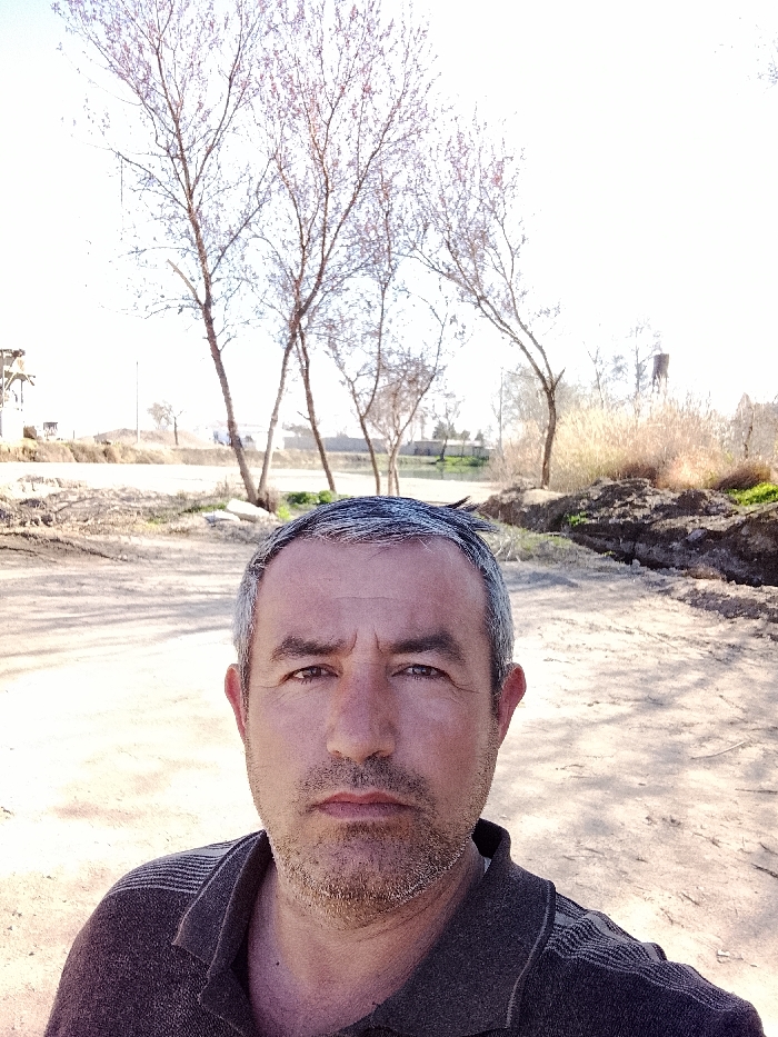 The image depicts a close-up selfie of a male subject in Yangi Chinoz, Uzbekistan, under a bright, clear sky. The man, appearing middle-aged, occupies the lower half of the frame. He has short, greying hair, a short beard, and dark eyes, looking directly into the camera with a neutral expression. He is wearing a dark-colored polo shirt with faint horizontal stripe patterns on the shoulders.
The background reveals an outdoor, unpaved environment. The immediate ground behind the subject is light-colored dirt or sand. Further back, several tall, slender deciduous trees, mostly bare with sparse reddish-purple buds, are visible. These trees border a narrow body of water, likely a canal, which reflects the bright sky. Beyond the water and trees, an unpaved road or pathway traverses the scene. To the right, mounds of excavated earth and patches of dry, tall grass or reeds are present. In the distant background, industrial or architectural elements are discernible: a tall, dark, cylindrical structure, possibly a water tower, on the far right, and a multi-story structure with what appears to be scaffolding or industrial equipment on the far left. Distant green vegetation and faint structures complete the horizon.