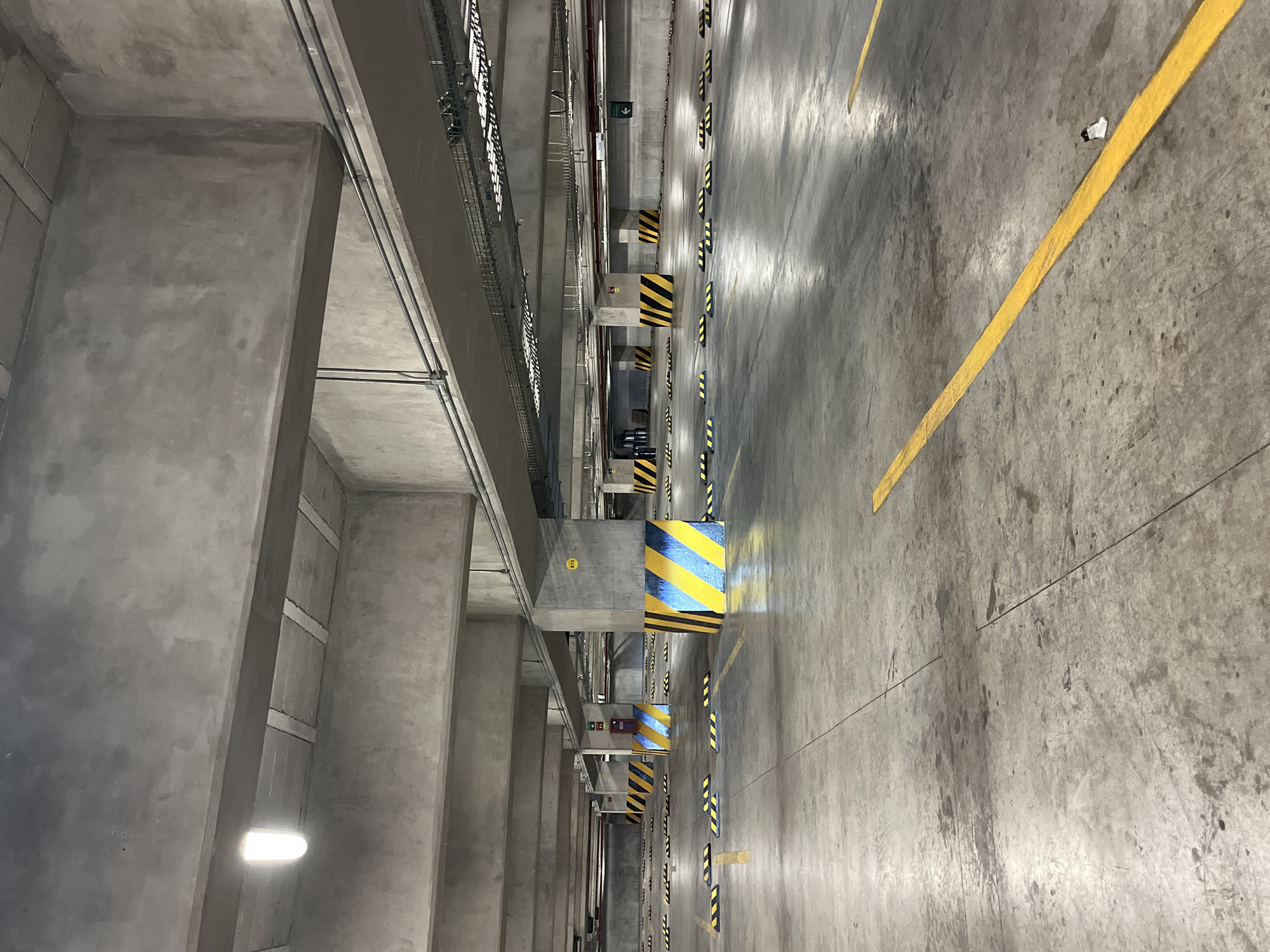The image shows the interior of a parking garage in Puebla, Mexico. The structure is primarily concrete, with pillars supporting the ceiling. Yellow lane markings are visible on the concrete floor, and pillars are marked with yellow and black hazard stripes. Electrical conduits and cable trays run along the ceiling. A few light fixtures are visible.