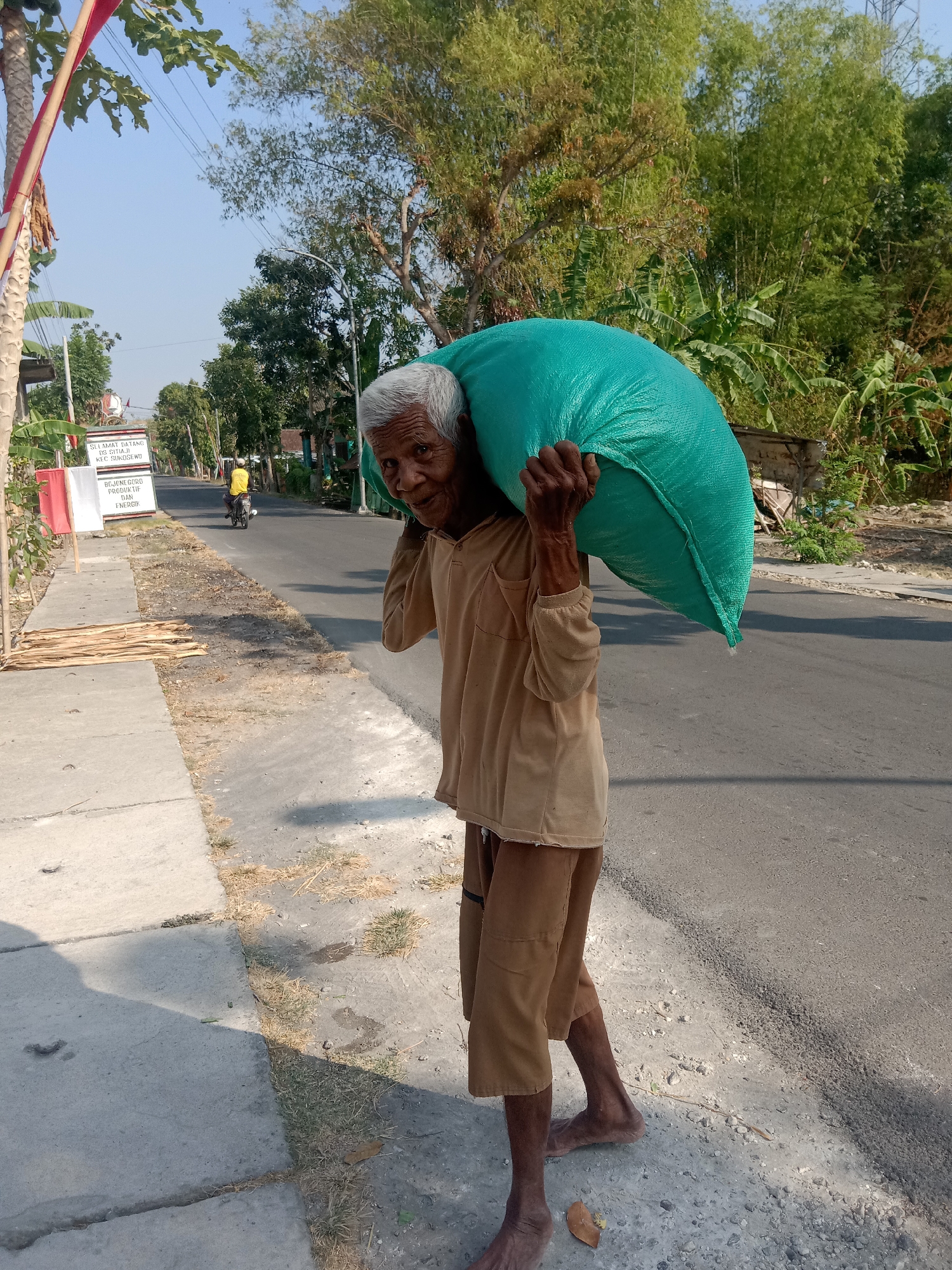 An elderly person with gray hair is walking on a paved road carrying a large, teal-colored sack on their right shoulder. The person is wearing a tan long-sleeved shirt and brown shorts. The person is barefoot. The scene is outdoors, with a sidewalk to the left of the road. There are trees and vegetation in the background, and a sign on the left side of the road that reads "SELAMAT DATANG DI SITIAJI KEC SUKOSEWU." This indicates the location is Sitiaji Village, Sukosewu District, in Indonesia. The road is asphalt, and there is some dry grass and small stones on the side of the road. A yellow motorcycle is visible in the distance on the road. The sky is clear and blue, suggesting it is daytime.