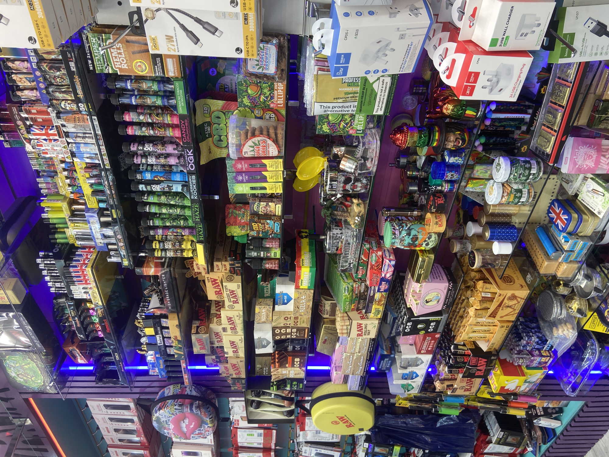 The image captures an extensive display of products within what appears to be a retail smoke shop or a convenience store specializing in related merchandise. No individuals are visible in the scene.
The store's interior features multiple shelves, many illuminated by blue LED strips, showcasing a diverse array of items. Products are densely packed, with prominent sections dedicated to smoking accessories. These include numerous lighters of various brands such as CLIPPER, Flamelock TORCH, and Bairui HIGH GRADE LIGHTER. A wide selection of rolling papers and tips is visible, with brands like RAW, RIZLA, OCB, ELEMENT, MARLEY, and Blazy Susan. Boxes of BROCONE cones and PINK CONES are also present.
A substantial portion of the display features CBD and hemp-related products. Brands like AZTEC CBD and DARWIN CBD are evident, alongside edibles such as SPACE BROWNIE, LOLIPOP, and items labeled "CANDY CRUNCHED," "BLACKBERRY OGK," "MYSTIKAL HAZE," and "KOOKIES OGK," some explicitly stating "CONTAINS NO TOBACCO OR NICOTINE." Also featured are "HEMPER HEMP ROLLS" with a "GLASS TIP" and "PROFESSOR HERB PREMIUM HEMP BLEND," some packaging carrying a warning: "This product is not a safe alternative to tobacco." An "18+" age restriction is visible on some product packaging.
Beyond smoking paraphernalia, the shelves also hold electronic accessories like USB-C TO USB-C and USB-C TO iPhone cables (ASPOR 27W, hoco.), and various chargers such as "3A USB Charging Plug" and "2USB WALL CHARGER" (Bitz PREMIUM, CP05-1 QC3.0). Other miscellaneous items include grinders, small containers, and decorative cases, one featuring a red lips graphic and another yellow case branded RAW. The overall impression is a vibrant, well-stocked retail environment with a focus on tobacco, vaping, and CBD-related goods.