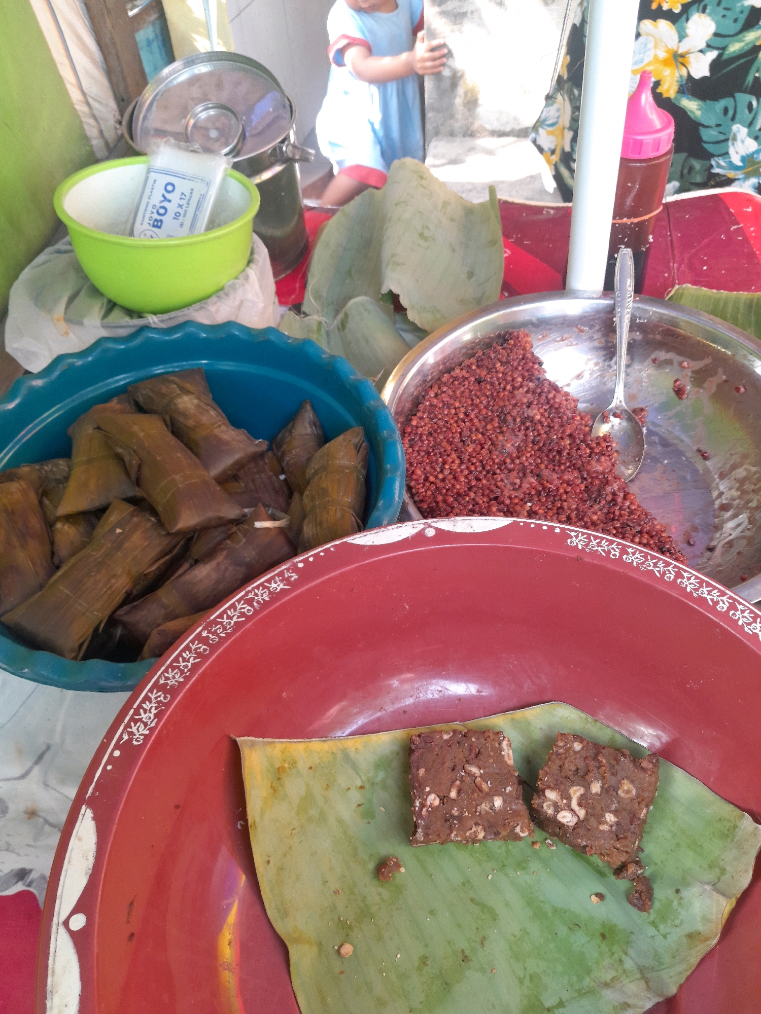 The image shows a close-up of a food stall in Jogosatru, Indonesia, with several food items displayed. In the foreground, two square pieces of a dark, dense confection, possibly a dessert, are placed on a banana leaf within a large red dish. To the left of the red dish is a blue bowl filled with numerous individually wrapped food items, appearing to be rice cakes or similar wrapped snacks. Behind this, a silver bowl contains a pile of small, reddish-brown grains, possibly a type of seed or grain, with a spoon resting in it. A bright green plastic bowl is visible to the left, containing a package of plastic bags labeled "BOYO 10 X 17". In the background, a child wearing a blue shirt and shorts is partially visible, holding onto a white pole. The overall environment suggests an outdoor market or street food vendor setting, with various textures and colors contributing to the scene.