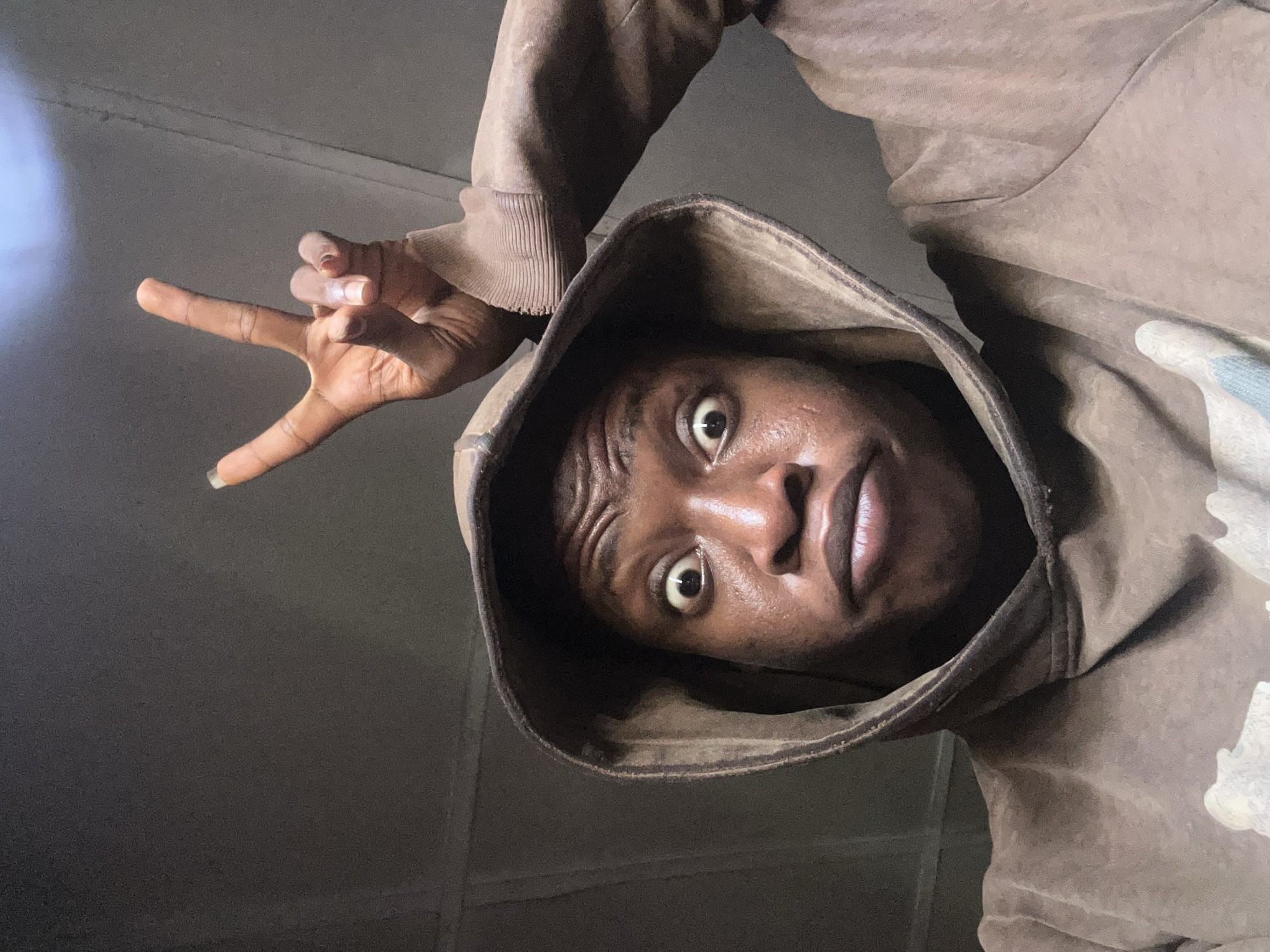 A low-angle close-up shows a person of African descent wearing a brown hooded sweatshirt. The person's face is prominently displayed within the hood, with wide eyes and a slightly furrowed brow. A hand making a "peace" or "V" sign is positioned to the left of the head. The background is dark and nondescript, with a patch of brighter light visible in the upper left. The location is Unknown, Unknown.