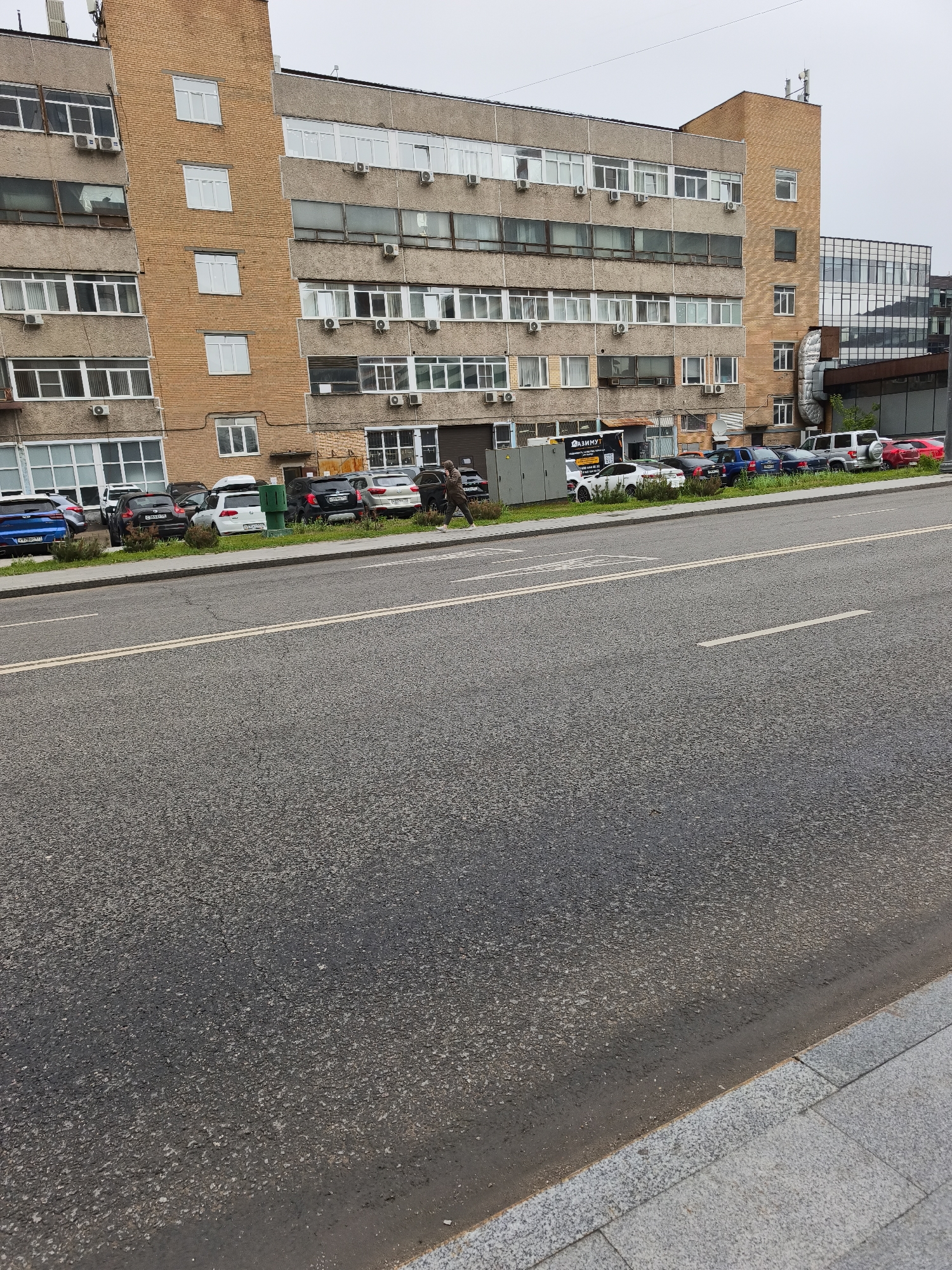 The image captures a street scene in Moscow, Russia. An asphalt road stretches across the foreground, with a sidewalk visible in the lower right corner. Parallel parked cars line the edge of the road adjacent to a multi-story, gray and beige-colored building. Air conditioning units are visible in the windows. Two people walk along the sidewalk in front of the building. In the background, additional modern glass and steel buildings rise behind the main structure. The sky appears overcast, lending a muted light to the scene.