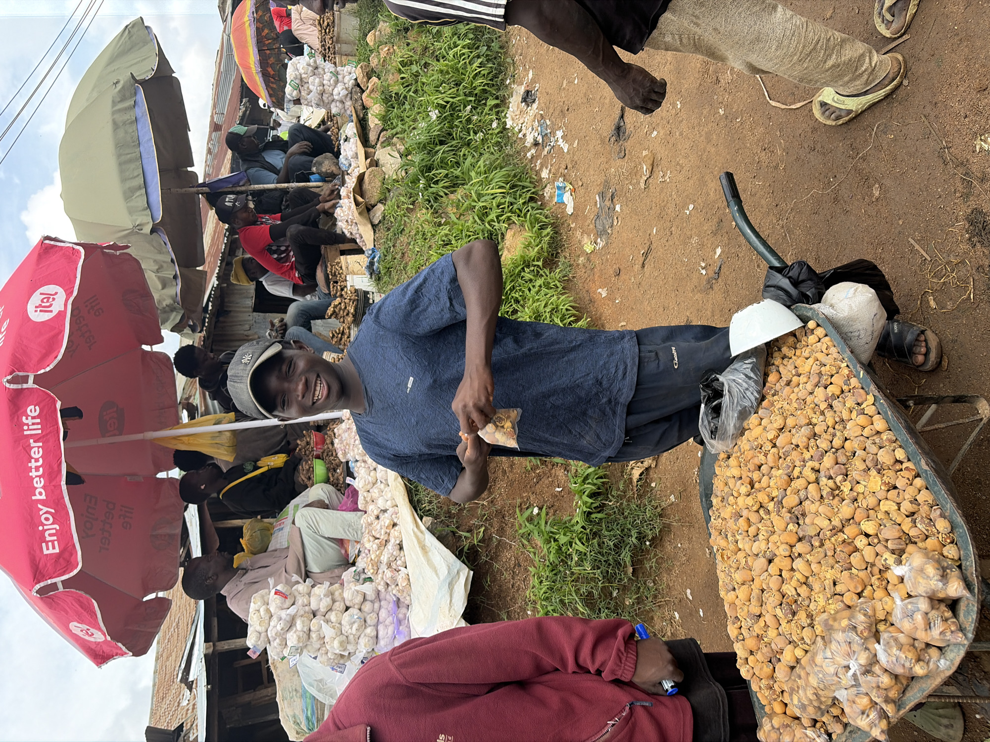 In Gura Top, Nigeria, a young man wearing a dark blue t-shirt and dark pants smiles at the camera, holding a small plastic bag containing food items. He stands in front of a wheelbarrow filled with a large quantity of small, round, yellow-brown food items, some of which are also bagged. The wheelbarrow is positioned on a dirt path in what appears to be an outdoor market. Behind him, several other people are visible, some sitting under red and green umbrellas, surrounded by various goods, including large piles of white bulbs, possibly garlic, and other produce. The ground is a mix of dirt and sparse green vegetation, with some scattered debris. The scene captures a moment of commerce and daily life in a bustling Nigerian marketplace.