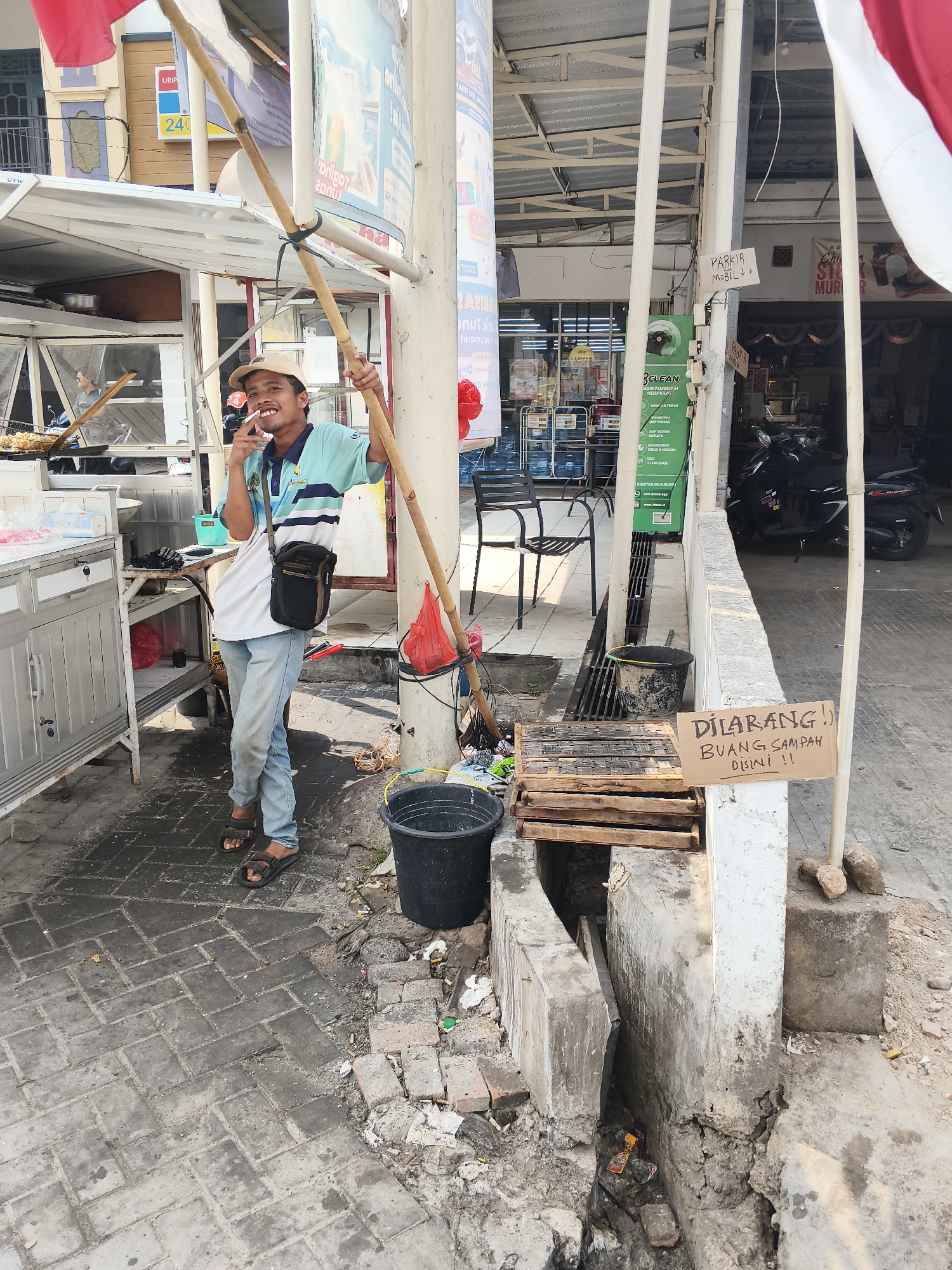A man stands in what appears to be an outdoor market or street-side stall area in Bandar Lampung, Indonesia. He is wearing a light blue and white striped polo shirt, light blue jeans, and dark sandals. A small black bag is slung across his body. He is holding a bamboo pole with both hands and has a cigarette in his mouth. He is looking towards the camera with a slight smile. Behind him, there is a food stall with a white awning and metal counters. To his right, there is a pole with several signs attached, including one that says "PARKIR MOBIL" which translates to "CAR PARKING". Further to the right, there is a sign on a cardboard piece that reads "DILARANG BUANG SAMPAH DI SINI!!" which means "DO NOT DUMP TRASH HERE!!". There are various objects scattered around the ground, including buckets, wooden pallets, and debris, suggesting a somewhat unkempt environment. A scooter is visible in the background. The ground is paved with interlocking grey bricks. The overall scene suggests a busy, informal commercial area.