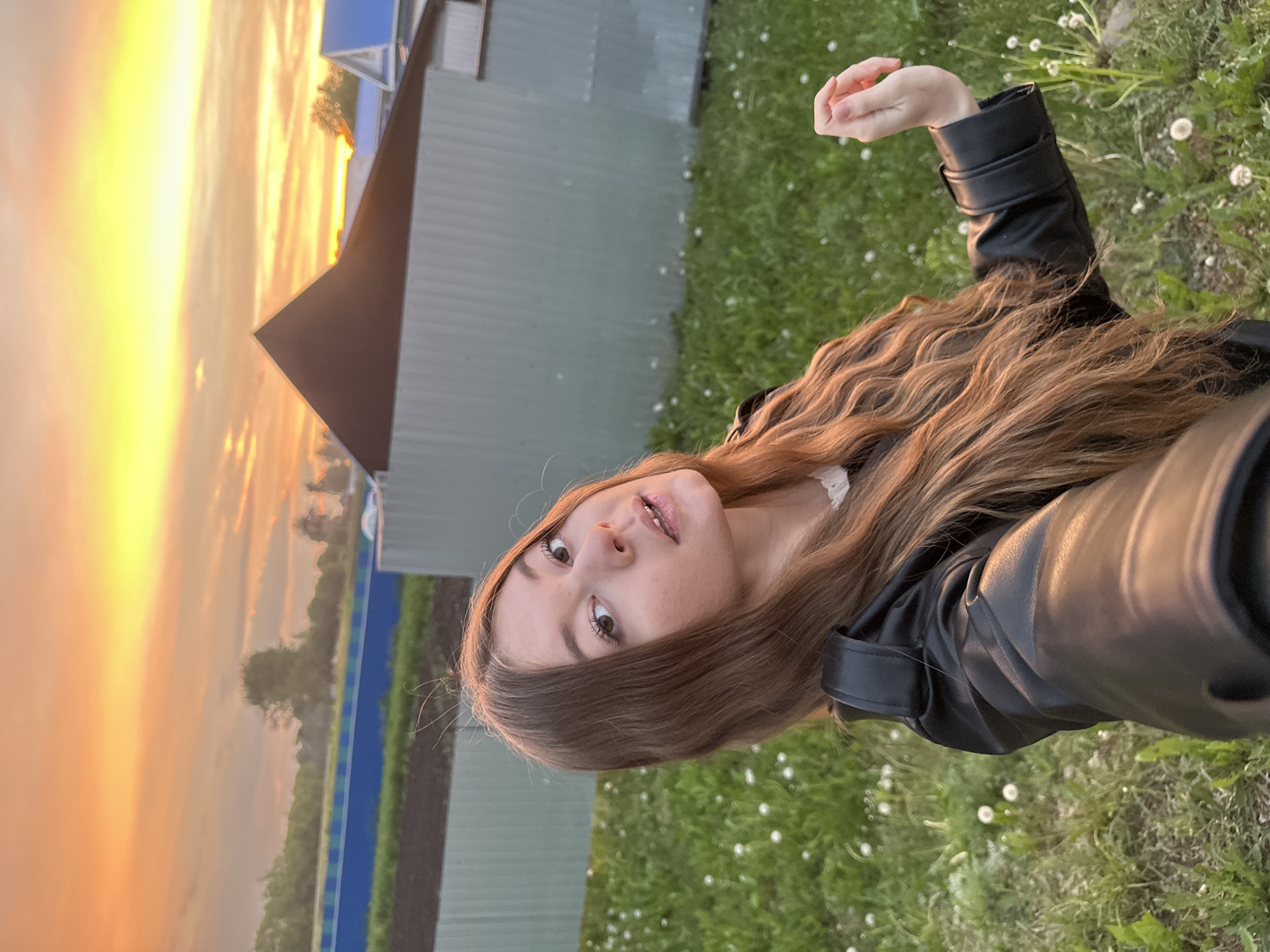 A young woman with long brown hair and wearing a black leather jacket is standing in a grassy area with dandelions. In the background, there is a metal fence with blue trim and a building with a brown roof. The sky is orange and yellow, suggesting a sunset or sunrise. The location is Кузембетьево, Russia.