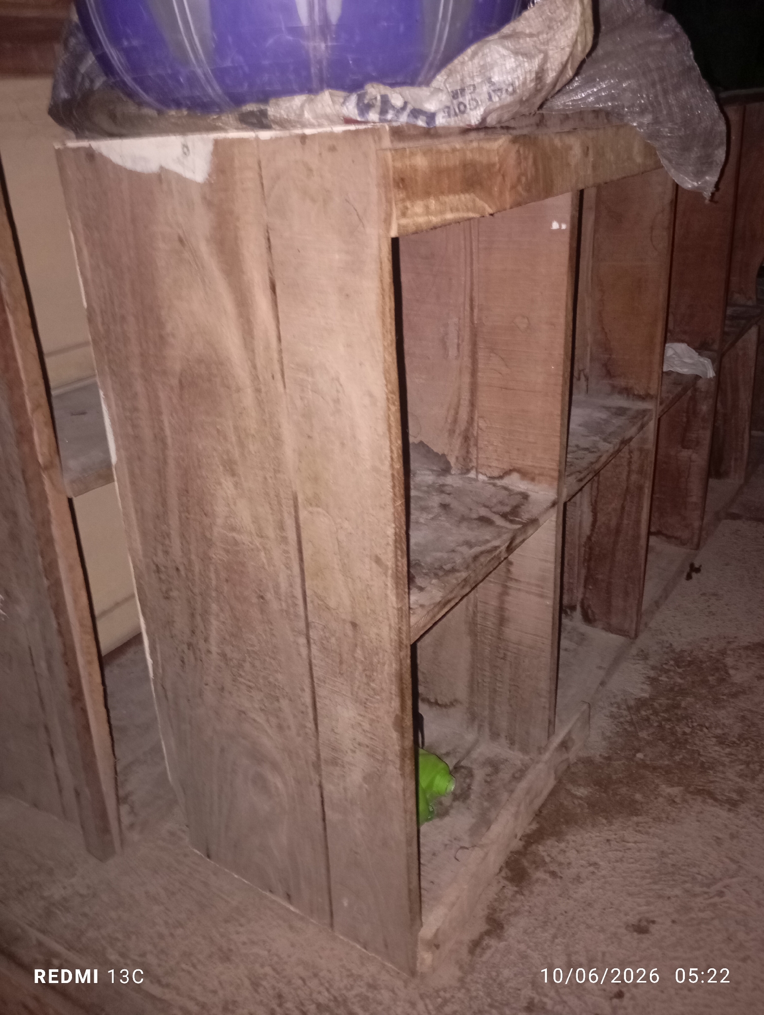 The image shows a roughly constructed wooden shelf unit. The shelves are exposed and appear to be dusty or possibly dirty. Some white material, possibly paint or primer, is visible on the top surface of the unit. A blue barrel or container is partially visible on top of the unit, covered with a woven sack and plastic sheeting. The scene is indoors, in what appears to be a storage area or workshop. The floor is uneven and dusty. The overall impression is one of a utilitarian and possibly temporary storage solution. The watermark in the lower right corner indicates the date and time are 10/06/2026 05:22 and the phone model is REDMI 13C. No specific location in Jos, Nigeria can be determined from the image itself.