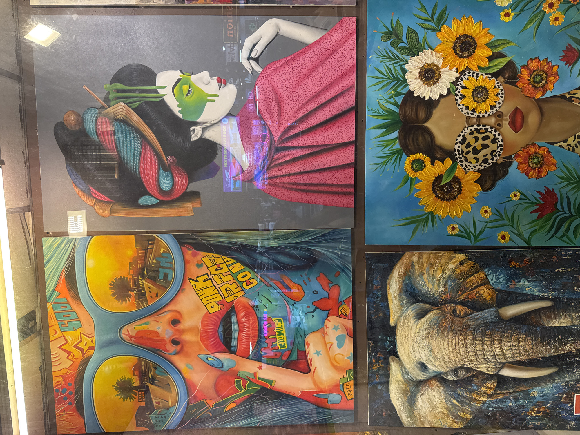 The image displays four distinct pieces of art, likely paintings, arranged in a grid. The context suggests they are on display in Patong, Thailand, possibly in a shop or market, as indicated by reflections of shop lights and a portion of a sign that reads "INFORMATION."
The top left artwork is a vibrant, close-up portrait of a face viewed through large, reflective blue sunglasses. The reflections in the sunglasses depict a cityscape with palm trees at sunset. The face is colorful, with a detailed mouth and a finger held to the lips, adorned with nail polish. Text, including "SOON" and "PUNK ROCK CONF," is incorporated into the artwork.
To the right of this is another artwork featuring a stylized portrait of a geisha-like figure. She has elaborate black hair with colorful, braided embellishments and is wearing a vibrant pink cloak. Her face is partially obscured by green paint around the eyes. Reflections in the artwork suggest the surrounding environment is a shop with signage, including reversed text that appears to be in English.
Below the geisha artwork, to the right, is a painting of a woman's face adorned with sunflowers and greenery as a headdress. She wears oversized, leopard-print sunglasses and flower-shaped earrings. The background is a bright blue.
The bottom left artwork is a detailed portrait of an elephant's head, rendered in a textured, impressionistic style with a dark, almost abstract background.
The overall scene captures a display of diverse artistic styles, hinting at a commercial environment in Patong, Thailand.