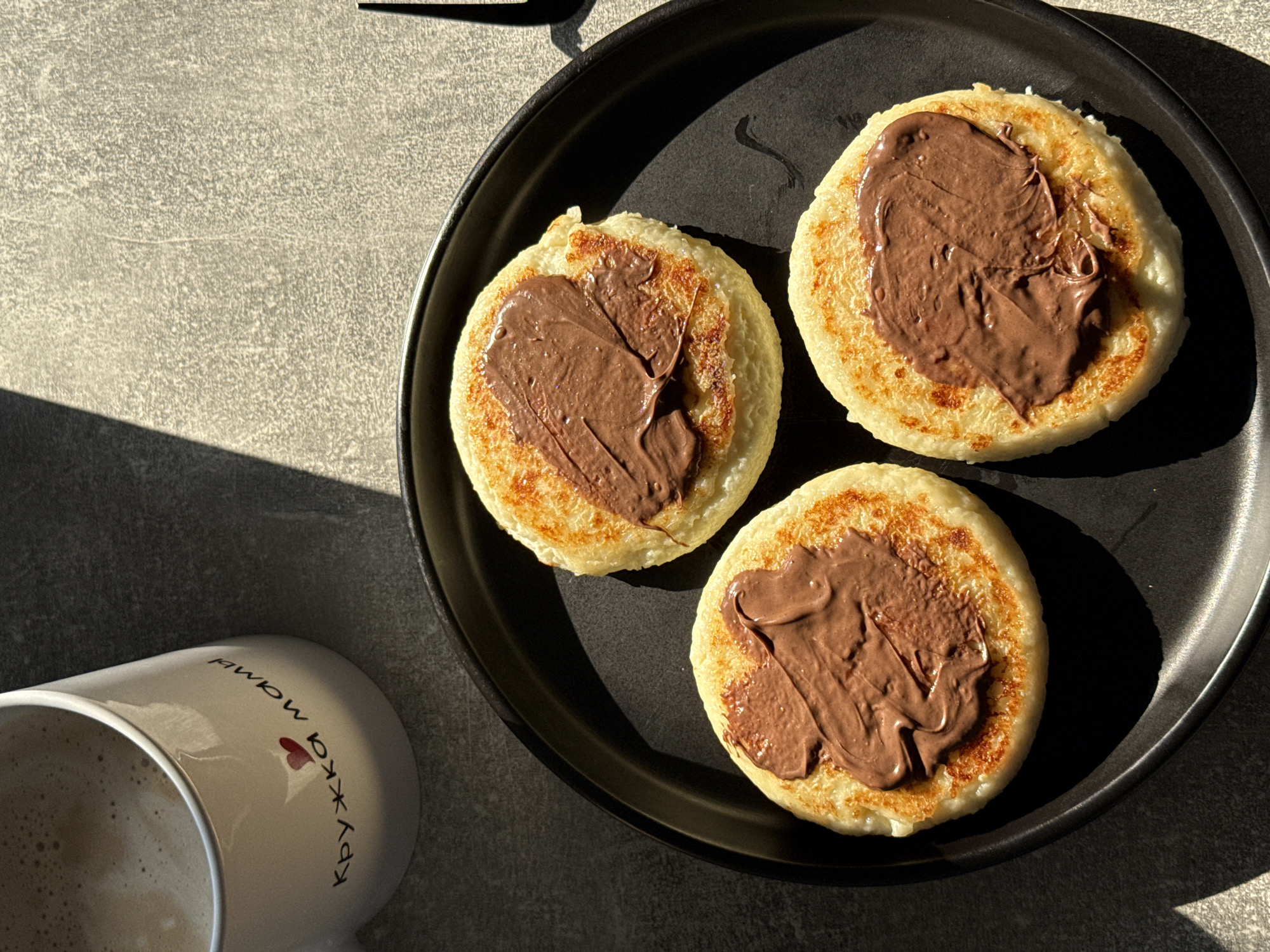A close-up, overhead shot shows three syrniki (a type of cheese pancake) topped with chocolate hazelnut spread arranged on a black plate. To the upper left of the plate is a white mug filled with coffee or a latte, which has text and a heart graphic on its side. The syrniki are golden brown and slightly uneven in shape, with a generous dollop of the chocolate spread on each. The mug has Cyrillic text which translates to "Mom's mug." The scene is set against a textured grey surface, likely a countertop or table. Harsh sunlight casts strong shadows across the scene, indicating a bright day, possibly late morning or early afternoon. The image does not provide any specific visual clues to pinpoint its location in Lyubertsy, Russia.