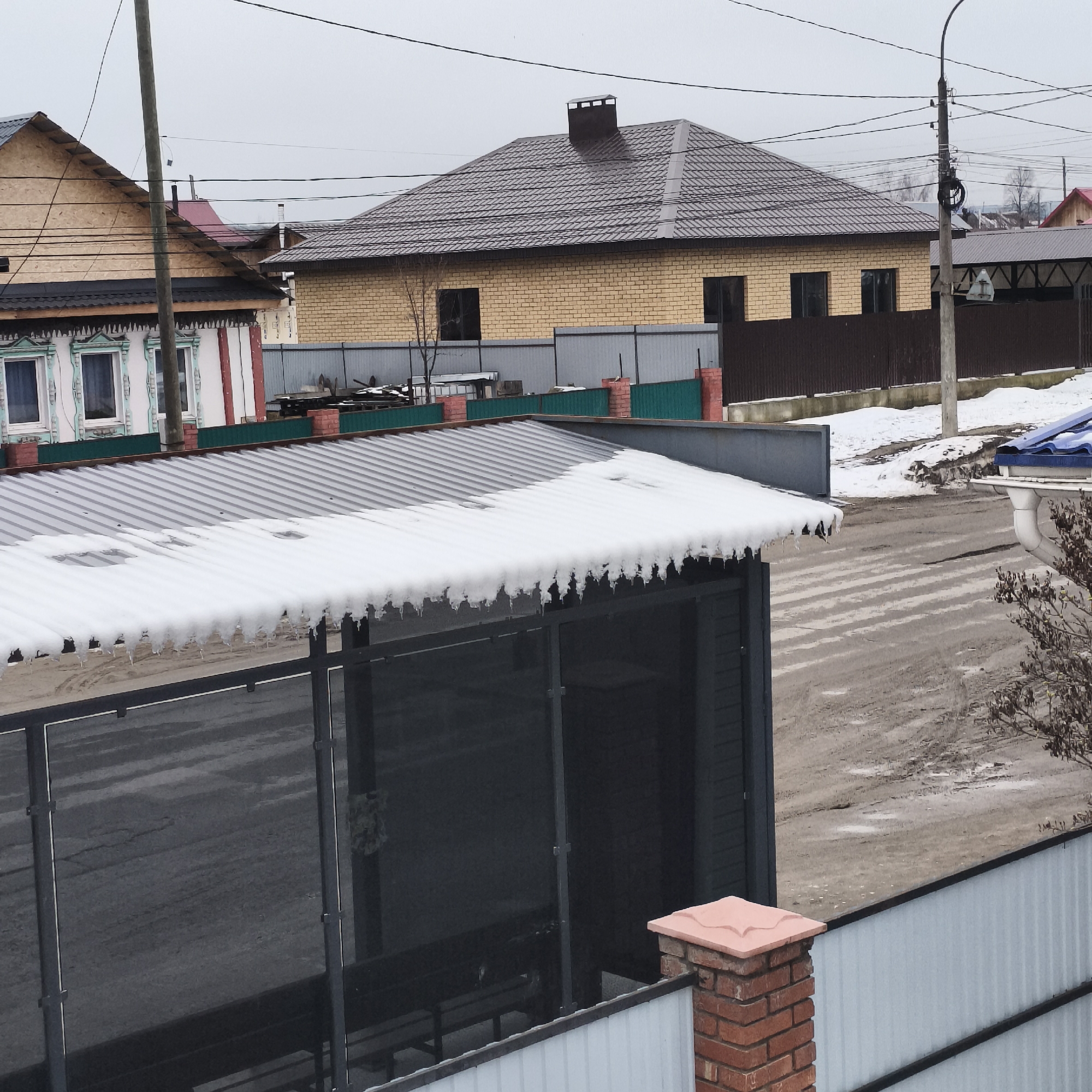 The image shows a residential street in Chaikovsky, Russia on an overcast day. The foreground features a corrugated metal roof partially covered in snow with icicles. Behind it is a dark, enclosed structure with a metal frame. In the lower right corner, a brick pillar topped with a decorative tile supports a white metal fence. The street appears wet and muddy with visible tire tracks. In the background, several houses are visible, including one with exposed wood, another with decorative trim, and a yellow brick house with a brown metal roof and chimney. Power lines stretch across the sky. Patches of snow are visible along the roadside.