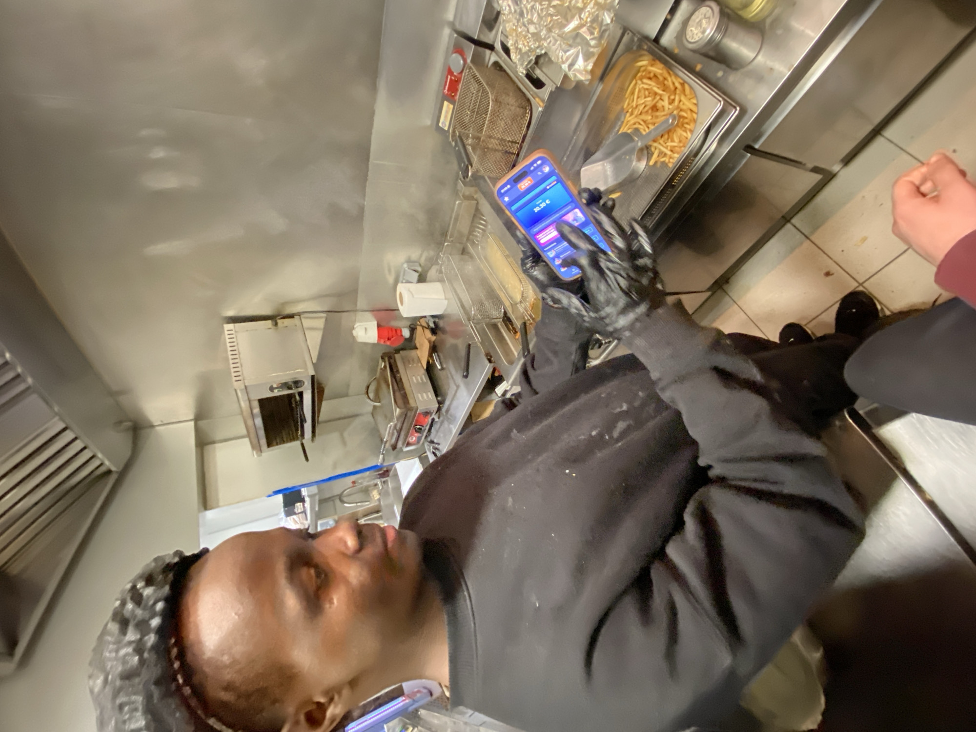 A person wearing black gloves and a black sweatshirt is looking at a smartphone while standing in a kitchen. A fryer and a container of french fries are visible on a stainless-steel counter. A second person's arm is partially visible in the lower right of the image. The kitchen appears to be in Schiltigheim, France.