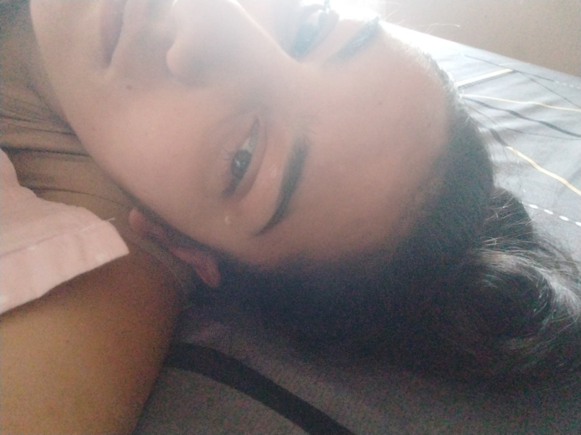 A close-up, slightly out-of-focus image shows a person lying on a bed. The person has dark hair and is partially facing the camera, with one eye visible. The person's skin is fair. A portion of a pink sleeve is visible on the left side of the frame. The bed has a gray and black striped pattern. The city and specific location are unknown.