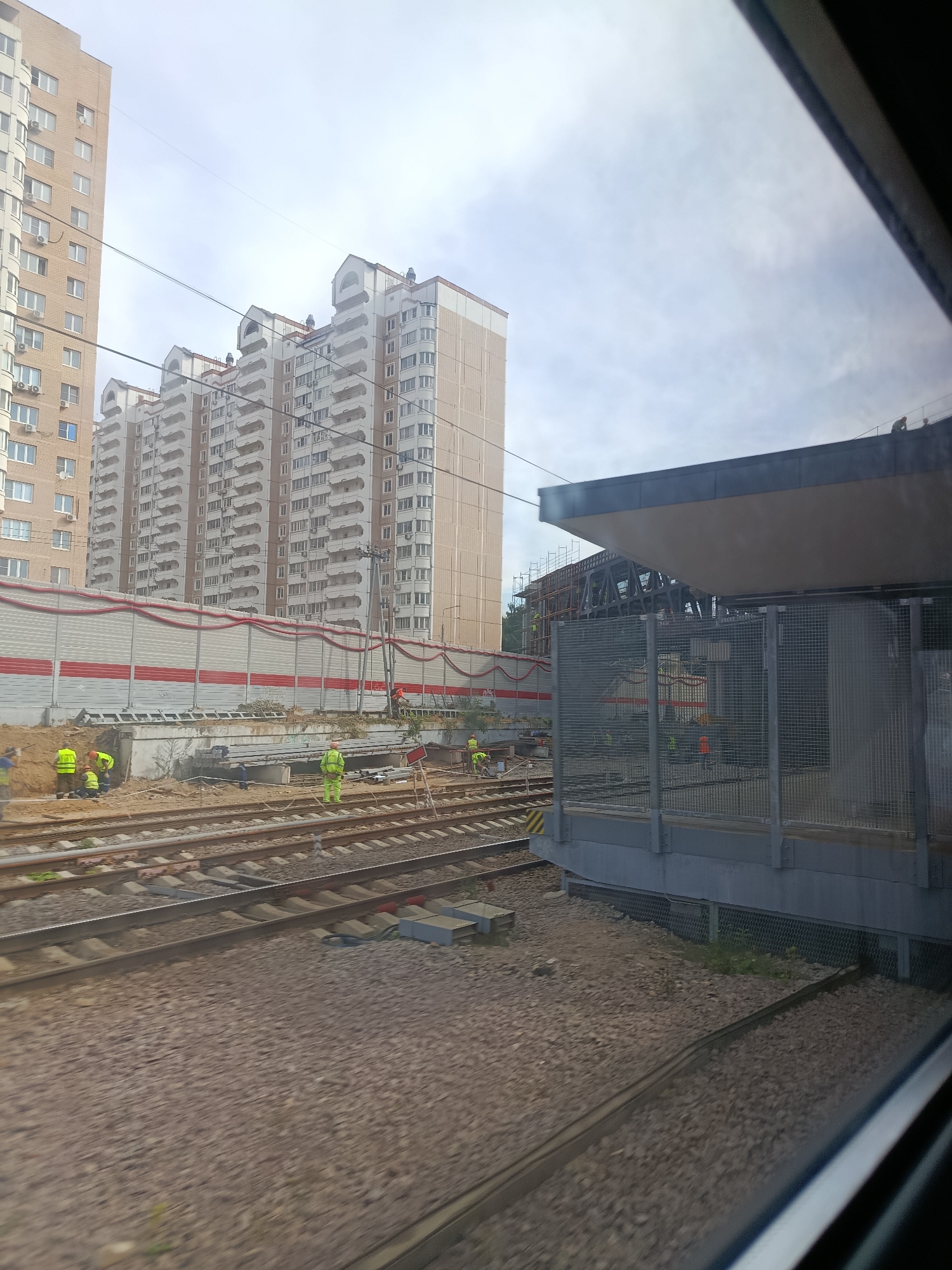 The image captures a construction site adjacent to railway tracks, likely in Balashikha, Russia, given the context. In the background, several tall, multi-story residential buildings with beige and white facades dominate the skyline. A prominent sound barrier, a long, light-colored wall with red wavy stripes, runs along the edge of the construction area. The construction site itself shows multiple workers in bright yellow vests and hard hats engaged in various tasks. Some are near the railway tracks, while others are further back, possibly working on foundations or structures. Equipment such as metal pipes and scaffolding is visible. In the foreground, the railway tracks are clearly laid out, with ballast stones beneath them. To the right, a modern structure, possibly a platform or station building with a gray roof and metal fencing, is under construction or has recently been completed. The sky is overcast, suggesting a cloudy day. The image appears to be taken from a moving vehicle, likely a train, due to the slight blur and the framing by a window.