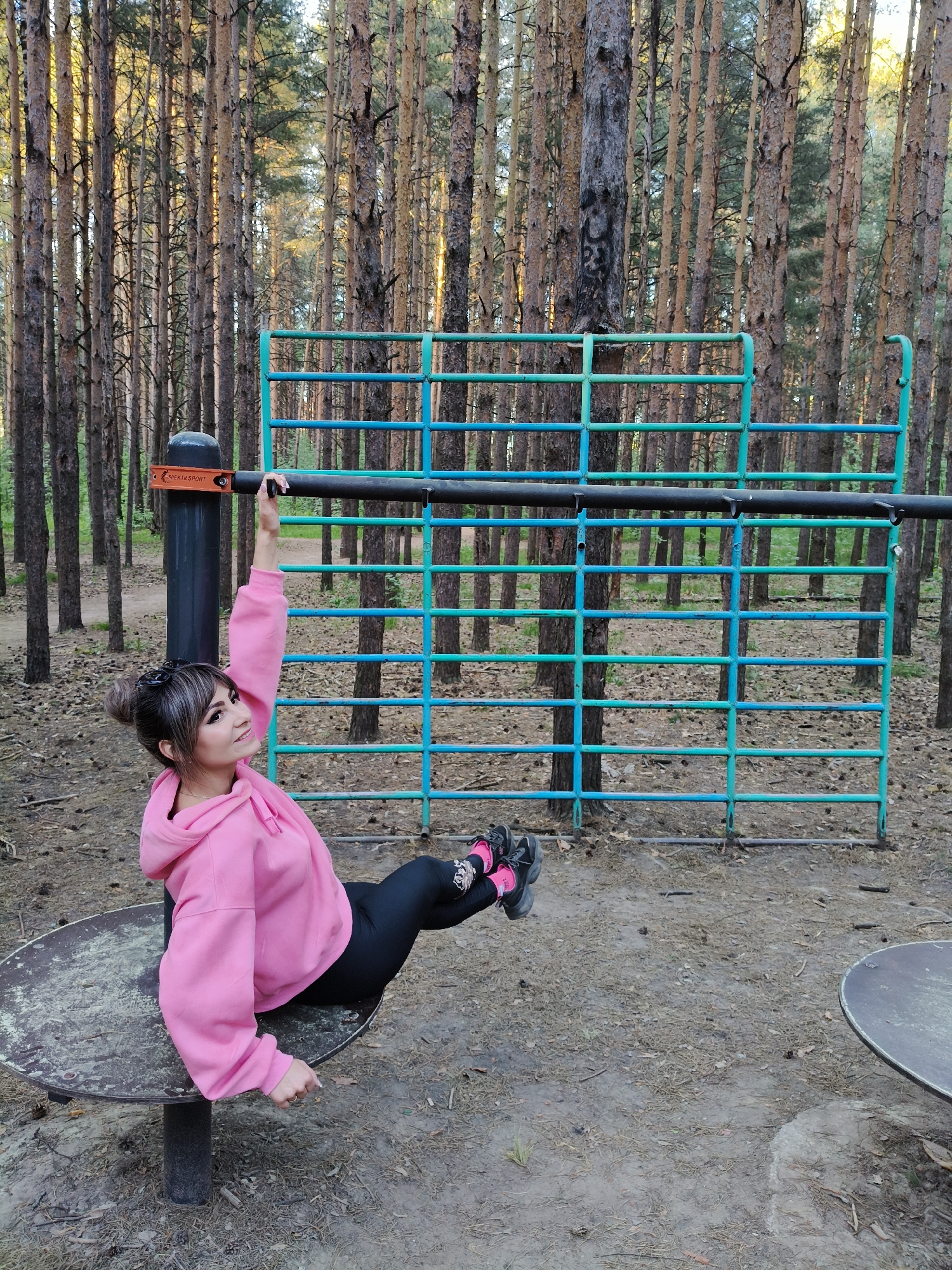The image shows a woman in a pink hoodie and black leggings performing an exercise in an outdoor workout area. She is holding onto a horizontal bar with one hand and sitting on a round, rotating platform. Behind her is a blue and green metal frame. The location appears to be a wooded area in Chaikovsky, Russia, based on the Cyrillic lettering on the workout equipment. The ground is covered with fallen leaves and dirt. There are tall, thin trees in the background.