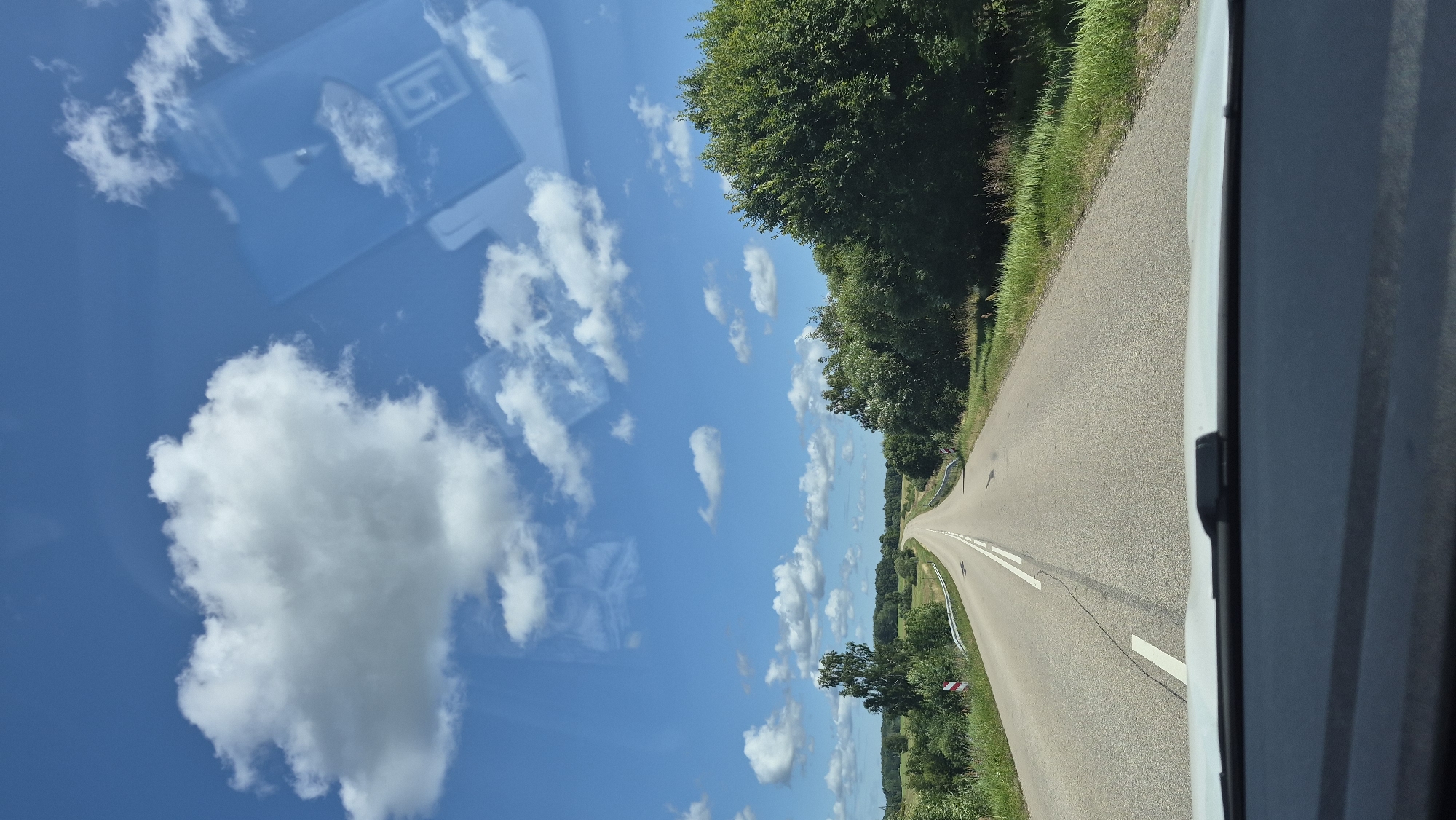 The image shows a view from inside a vehicle traveling on a paved road in Lučķini, Latvia. The road is lined with green grass and trees on both sides, and extends into the distance, curving slightly to the left. The sky is blue with scattered white clouds. The dashboard of the vehicle is visible in the foreground.