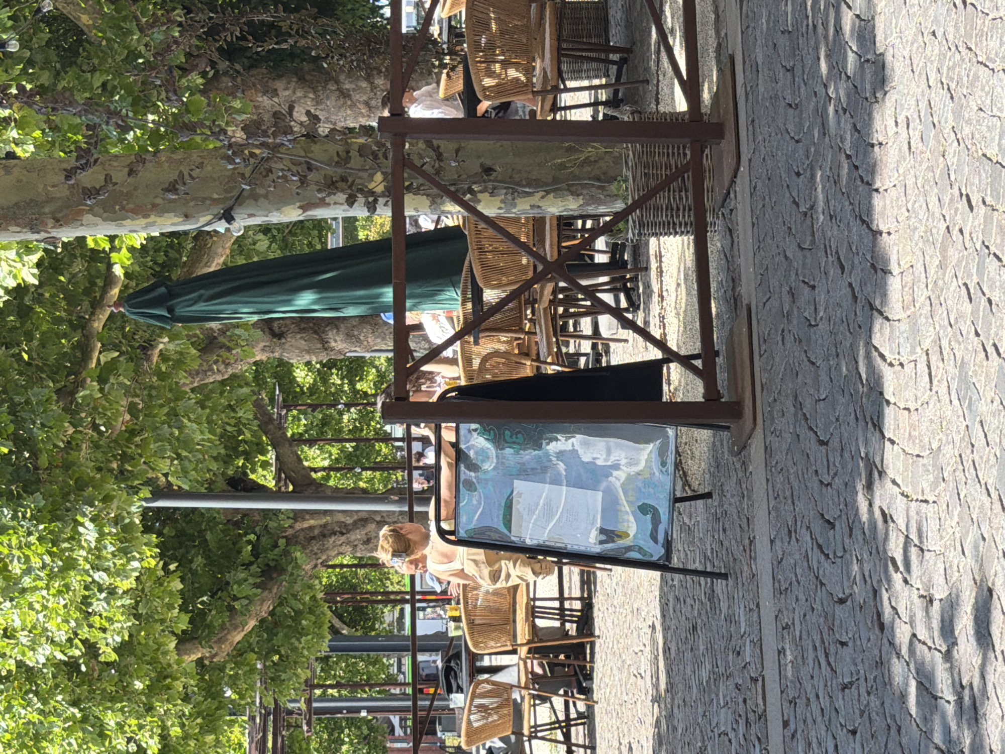 The image depicts an outdoor seating area of a cafe or restaurant, likely in Lund, Sweden. The foreground is dominated by a cobblestone pavement, with dappled sunlight creating shadows. A black A-frame sign with a colorful, illegible menu is prominently positioned. Behind the sign, several wicker chairs surround dark-colored tables under the shade of large trees with leafy canopies. A green umbrella, closed and leaning against a tree trunk, is visible. In the middle ground, several patrons are seated at tables, one woman with blonde hair pulled back is looking away from the camera. The background shows more trees and foliage, contributing to a relaxed, outdoor dining ambiance. The overall scene suggests a pleasant day, with people enjoying refreshments outdoors.