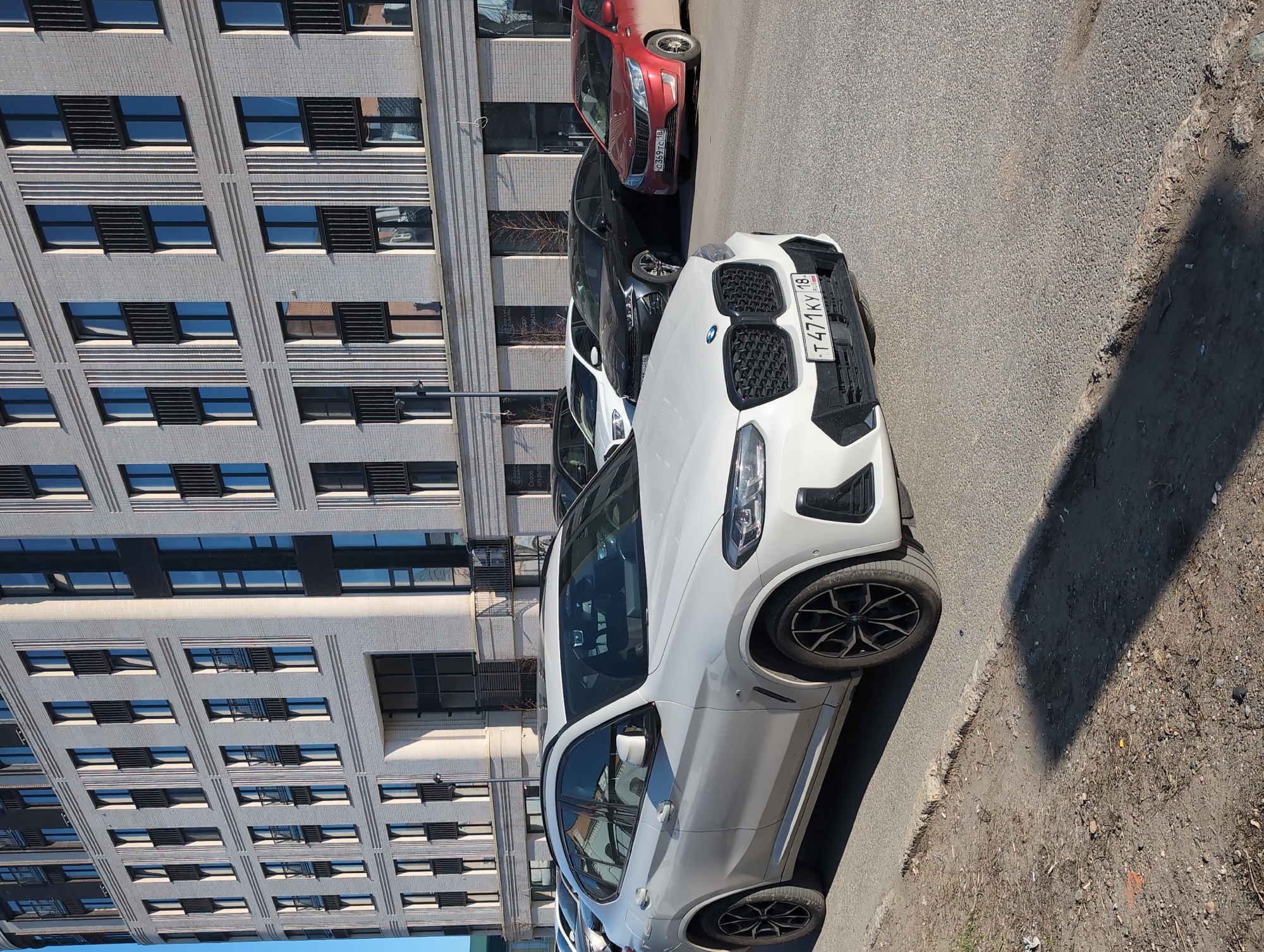 The image shows a white BMW X4 M parked on the side of a street in Izhevsk, Russia. The license plate number is T 471 KY 18. The building to the left is a multi-story building with many windows. Several other cars are parked behind the BMW, including a red Kia. The street surface is asphalt, with a rough edge and some dirt and debris on the side. The time of day appears to be daytime, with sunlight casting shadows on the road.