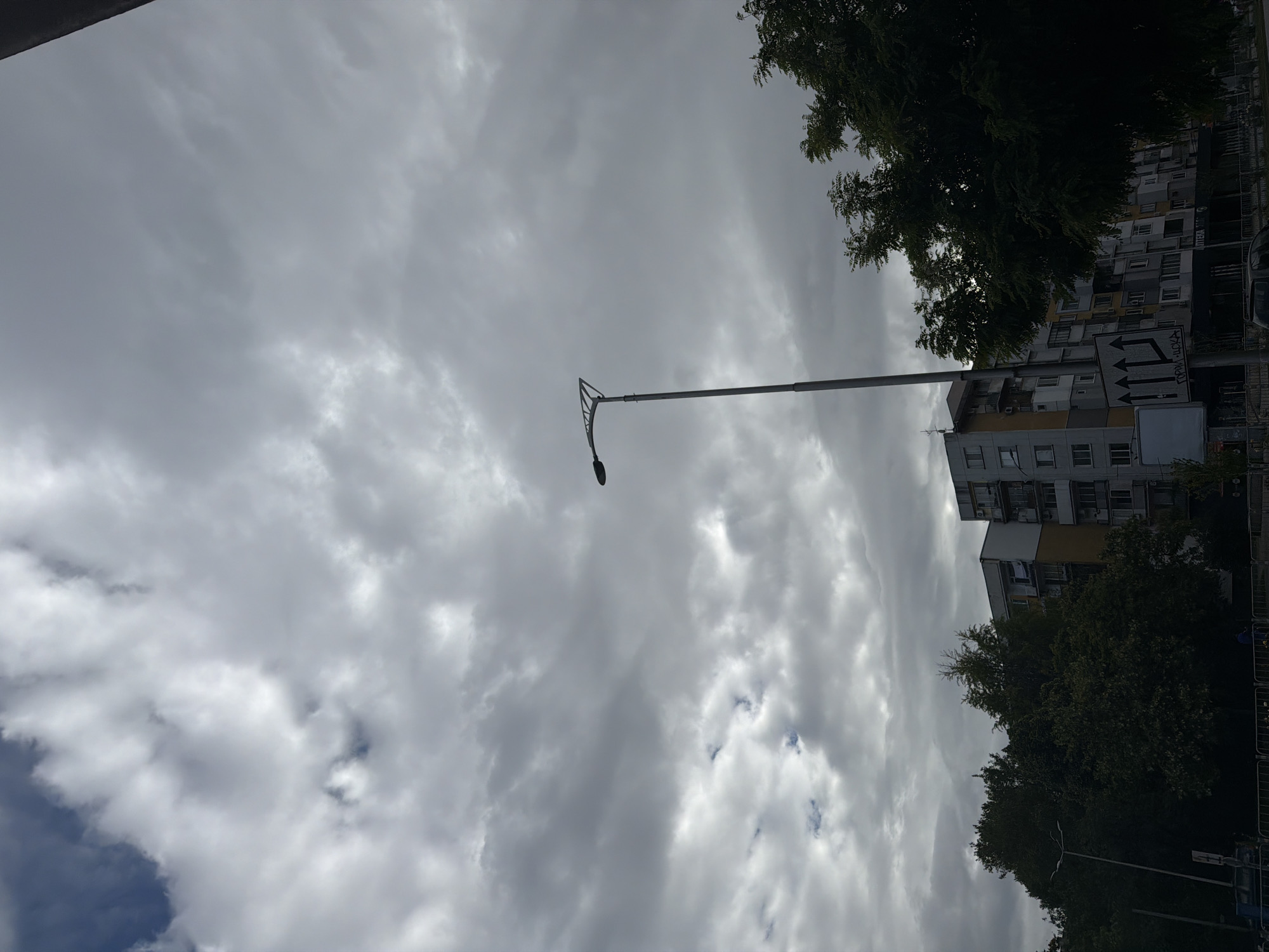 The image shows a view looking up towards a cloudy sky in Plovdiv, Bulgaria. In the foreground, there is a tall street lamp with a single light fixture. To the right, a multi-story apartment building is partially visible, obscured by trees. A signpost with directional arrows points in multiple directions, indicating street names or zones written in Cyrillic script. The overall atmosphere suggests an overcast day.