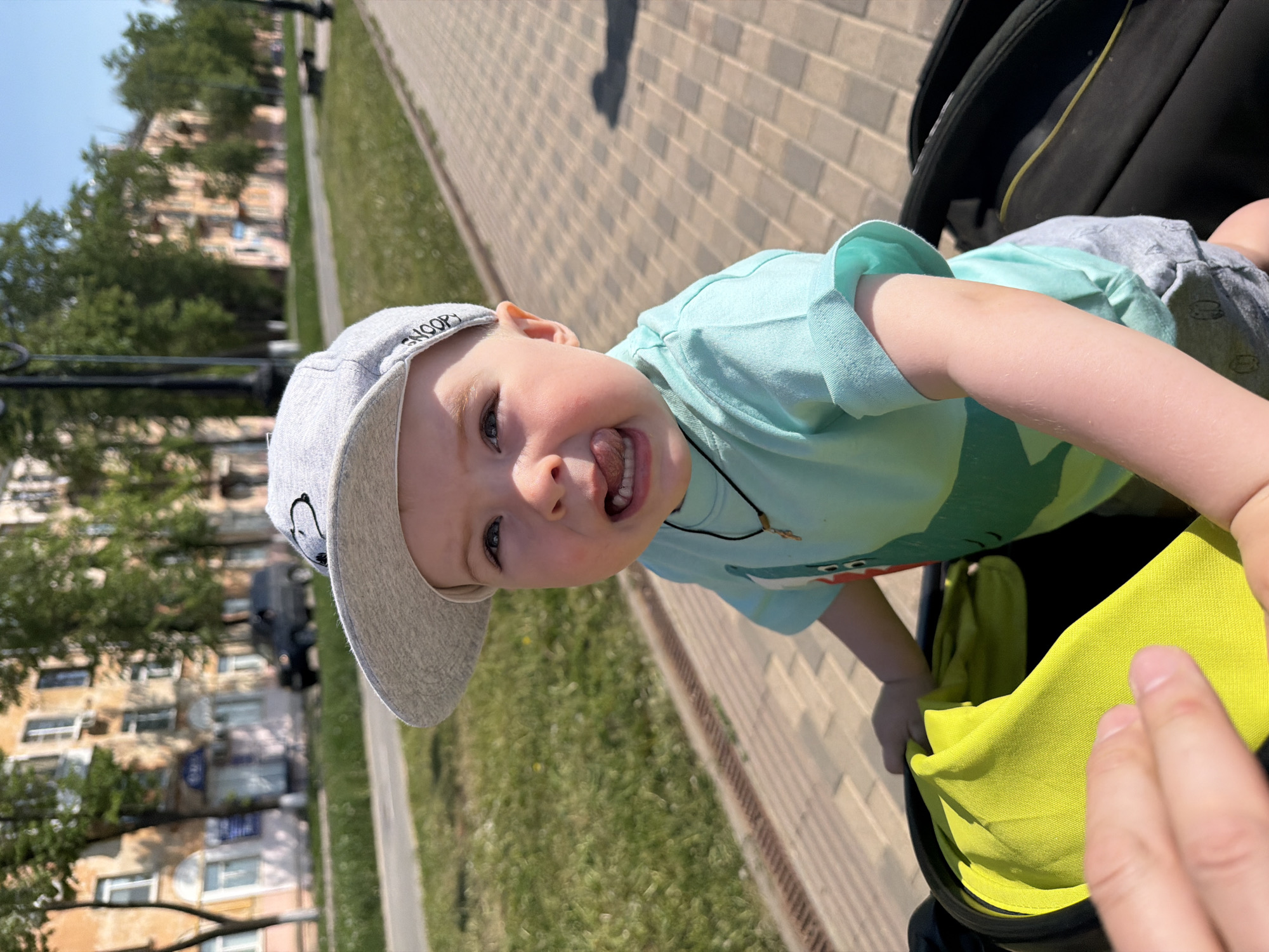 A young child wearing a light gray baseball cap with a cartoon character on it and a light blue T-shirt with a green pattern is seated in a stroller. The child has fair skin, blue eyes, and is sticking out their tongue. The stroller has a black frame and a bright yellow sunshade. A hand is visible in the lower right corner of the frame.
The background reveals an urban environment. A sidewalk made of light-colored bricks is next to a strip of green grass. Behind the grass, buildings are visible with trees partially obscuring them. A black car is parked on the street. This is Votkinsk, Russia.