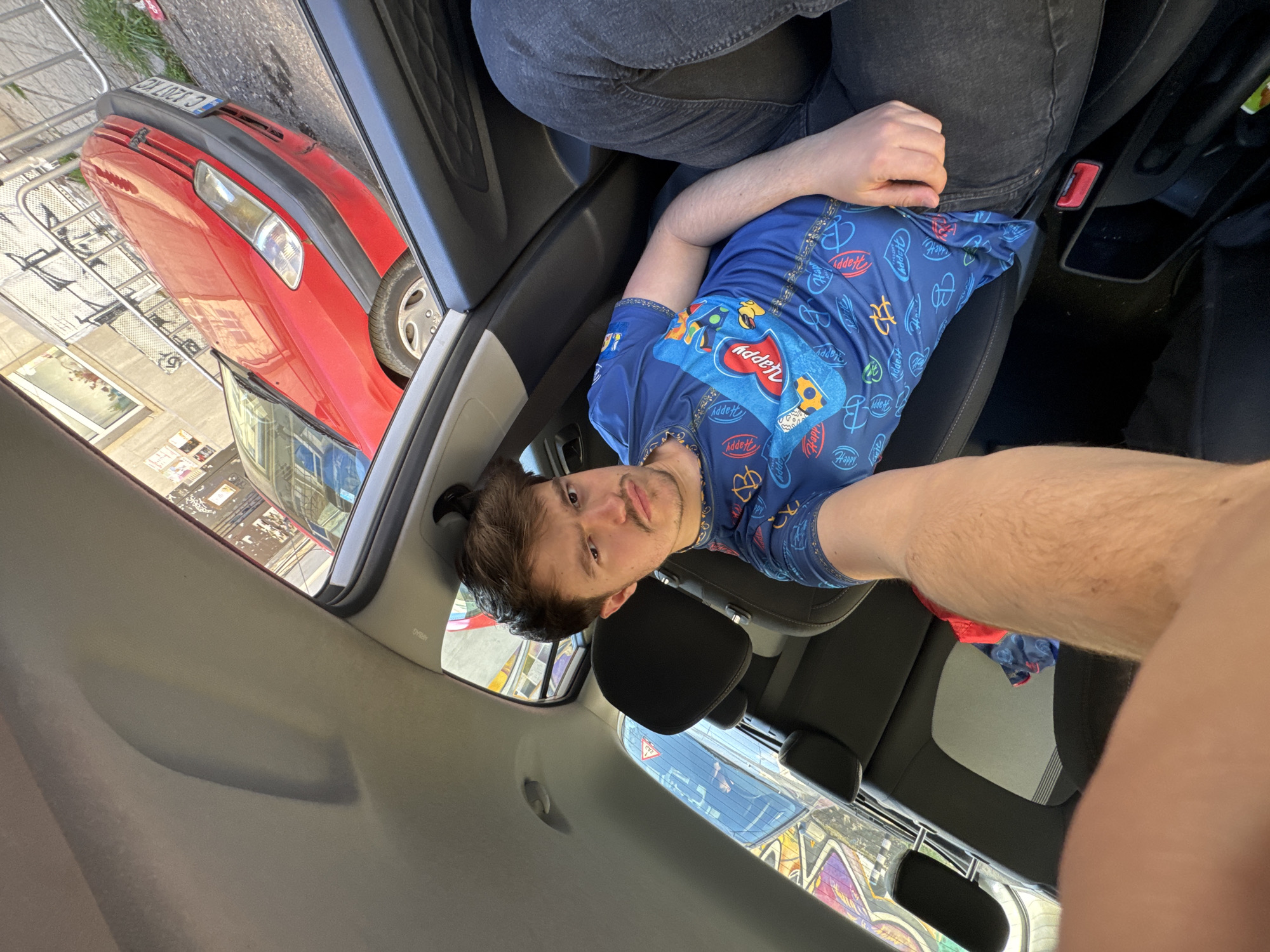 A medium shot shows a man wearing a blue patterned shirt inside a car. The car is parked in Sofia, Bulgaria, as evidenced by the license plate "C 2107 CO" on a red Peugeot visible through the car window. The man appears to be in the passenger seat. A second person is visible beside him, wearing jeans, but is out of focus. The environment outside the car includes other vehicles and urban architecture.
