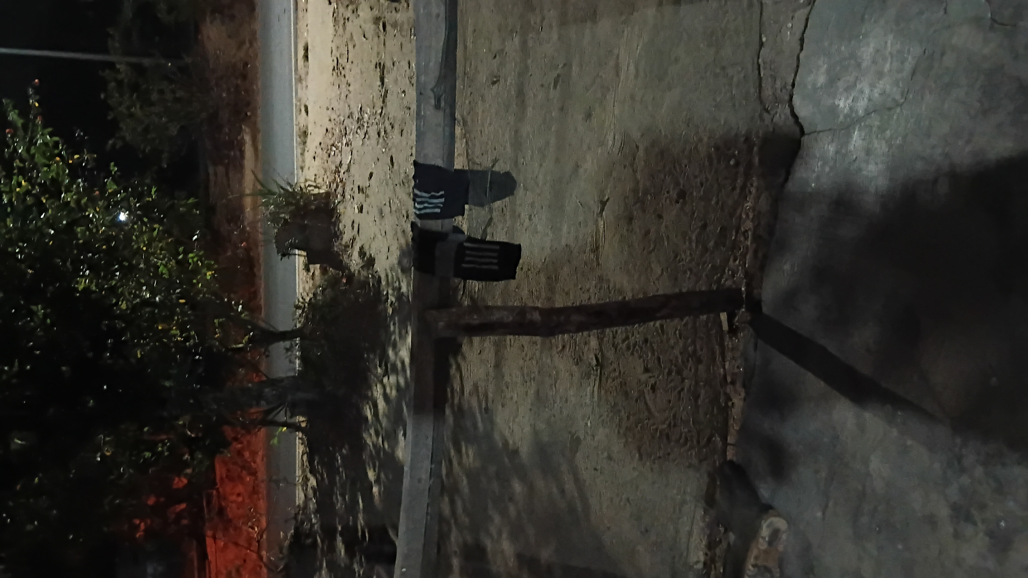 The image shows a dimly lit outdoor scene, likely at night, with a focus on a wooden beam supported by a vertical post. Two pairs of socks are hanging from the beam. One pair is black with white stripes, and the other is dark blue with white stripes. The ground beneath the beam appears to be concrete or packed dirt, with shadows cast by the beam and post. In the background, a tree with dark foliage is visible, and beyond that, a road or pathway. The overall impression is one of a simple, possibly rural or semi-urban environment in Prabumulih, Indonesia. There are no people or identifiable buildings directly in the frame, though a faint reddish glow in the far background might indicate artificial lighting.
