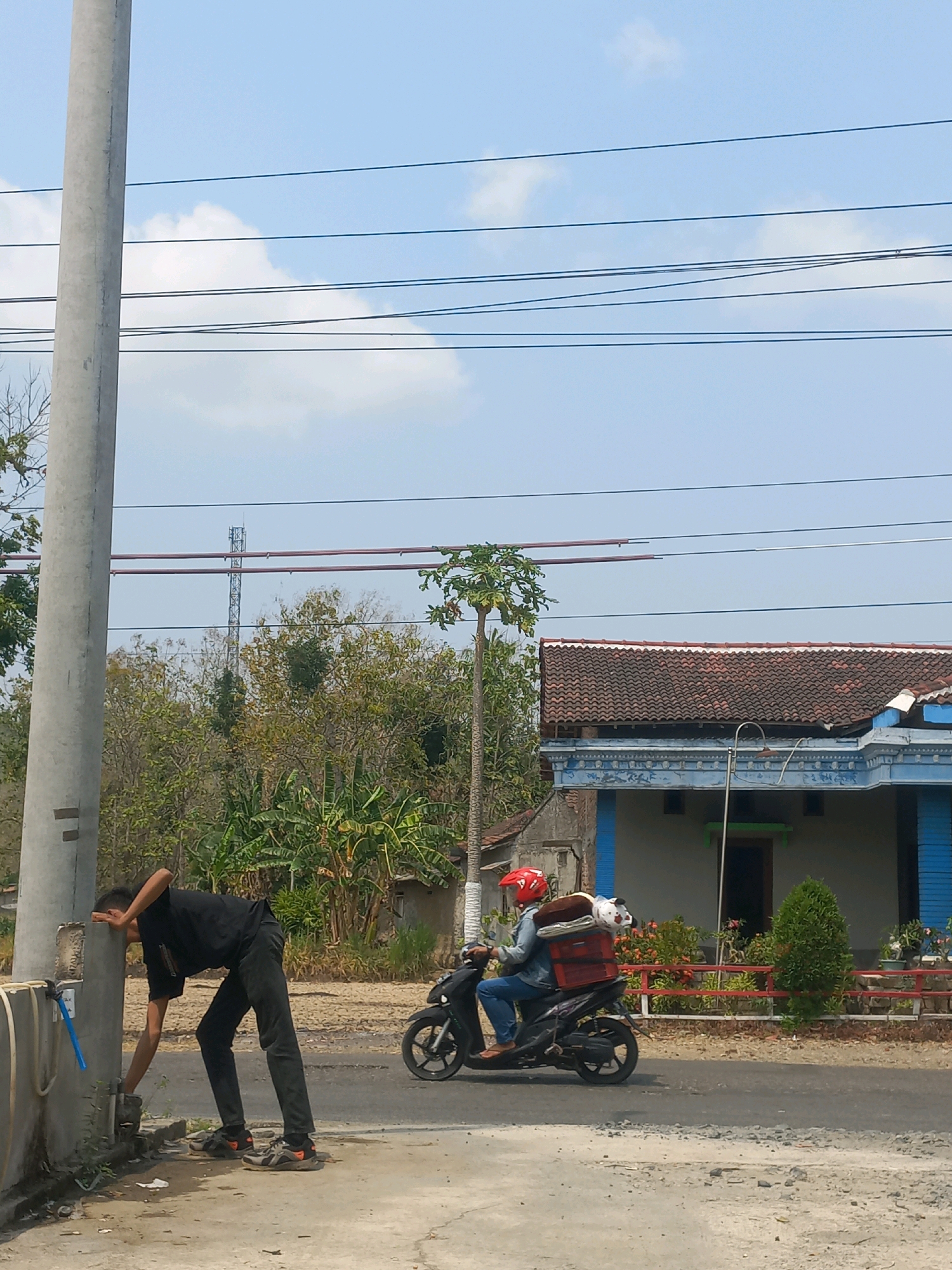 The image depicts an outdoor scene in Baturetno, Indonesia. On the left, a person in a black t-shirt and black pants is bent over, interacting with a water pipe or faucet attached to a concrete pole. They are wearing gloves and appear to be working on or around the pipe. To the right, a person on a scooter is riding past. This rider is wearing a red helmet, a denim jacket, and blue jeans, and is carrying a red crate on the back of the scooter, which contains several items. In the background, a house with a red tile roof and blue trim is visible, along with trees and vegetation. Power lines stretch across the sky, and a communication tower can be seen in the distance. The ground is paved with concrete and gravel. The overall lighting suggests a bright, sunny day with a clear blue sky and some scattered clouds.