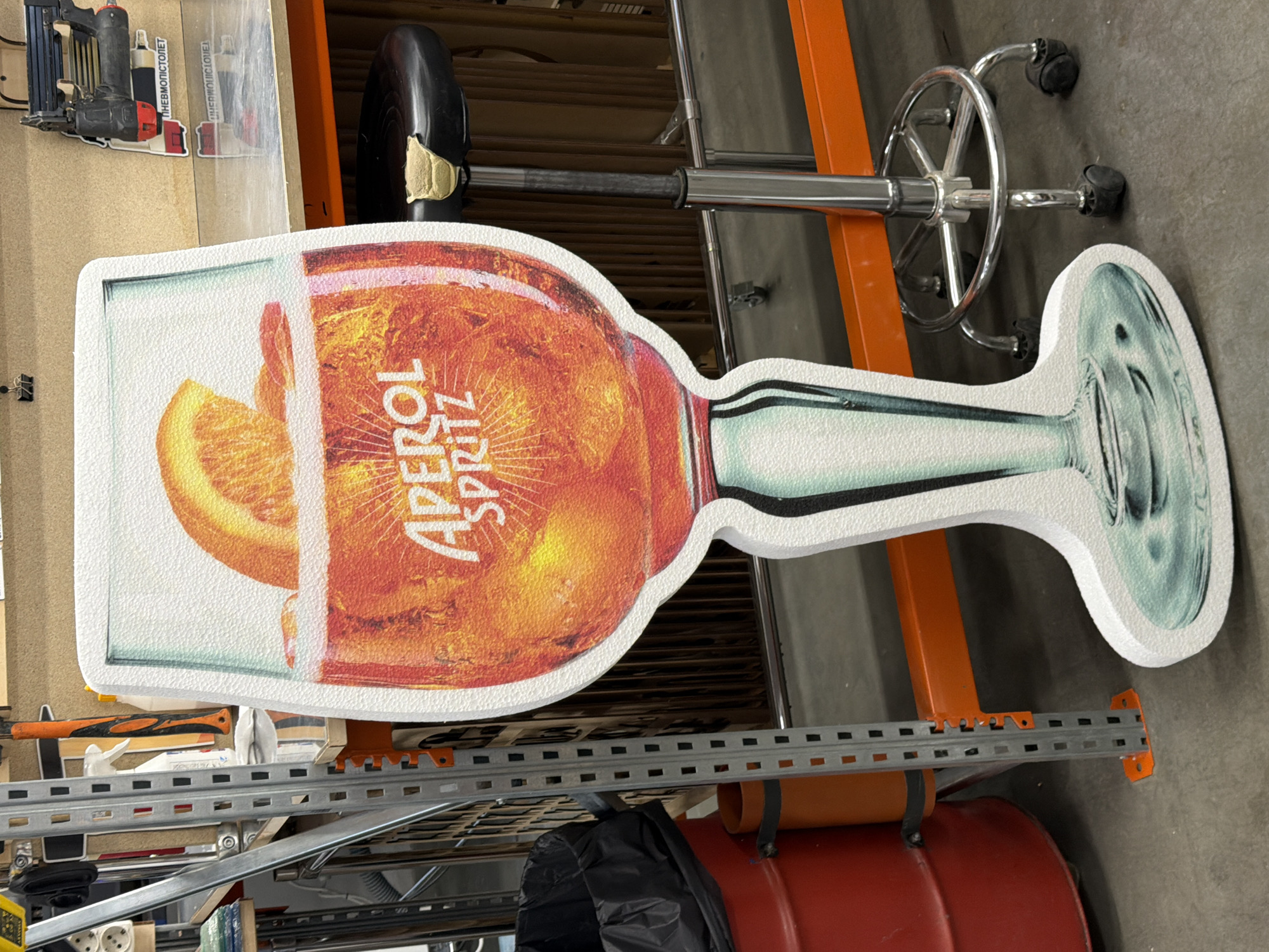 The image shows a life-size cutout of an Aperol Spritz cocktail glass placed in what appears to be a workshop or storage area. The cutout is white-bordered and features a detailed illustration of the orange-colored drink with ice and an orange slice. The background includes industrial shelving units, tools, a rolling stool with a black seat and chrome base, a red barrel, and a black trash bag. The overall environment suggests an indoor, possibly commercial or industrial, setting. There is no visible indication of the location being in Ternopil, Ukraine.