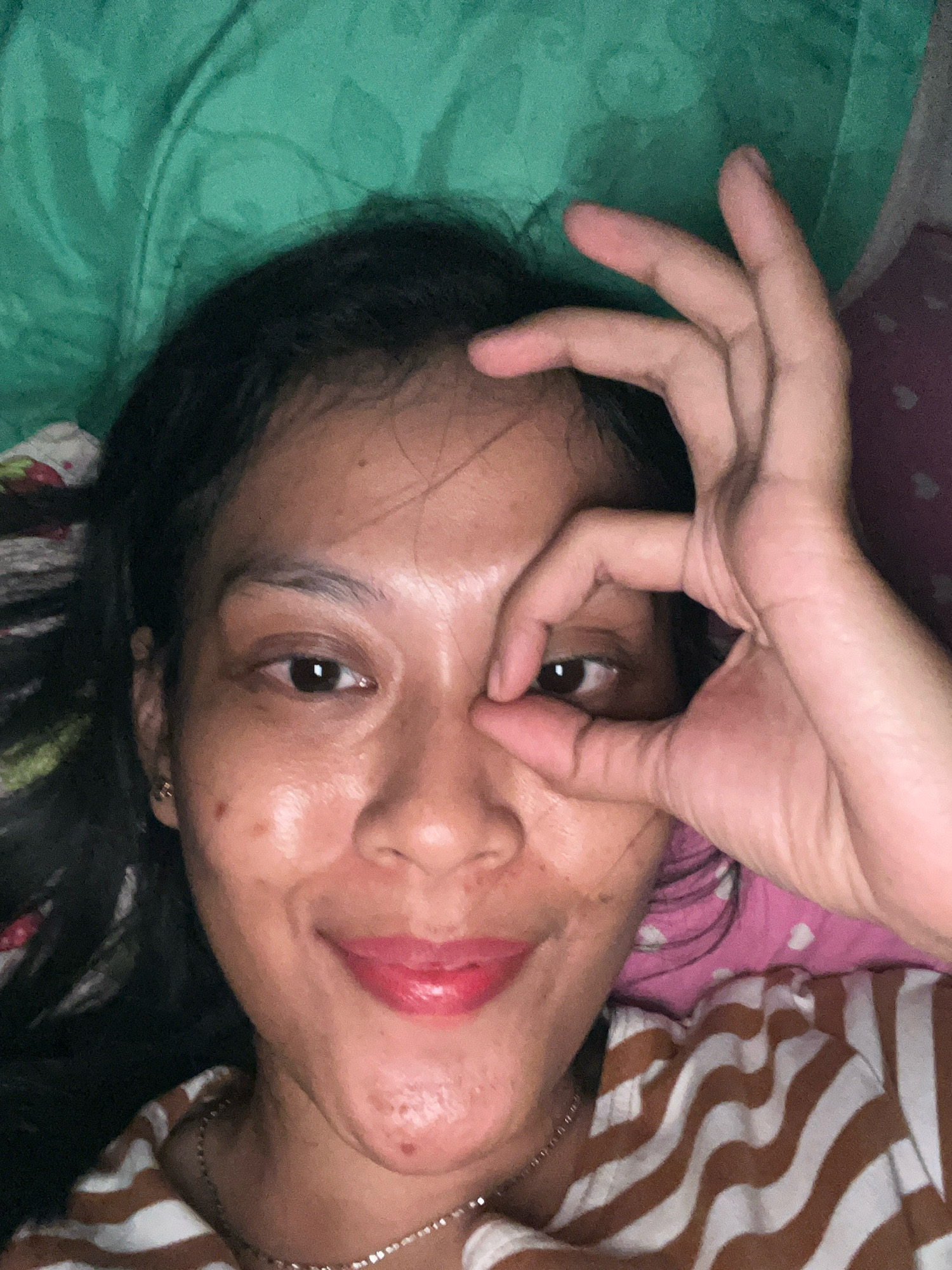 In a close-up, overhead shot, a woman of East Asian descent lies on a bed. She has dark hair, brown eyes, and fair skin with some blemishes. Her lips are painted a bright pink. She is wearing a brown and white striped t-shirt and a silver necklace. Her right hand is raised, with her thumb and index finger forming a circle around her right eye, while her other fingers are extended upwards. The background consists of a green blanket and what appears to be pink fabric with white hearts. The image is lit by an unseen light source, creating a slightly grainy texture.