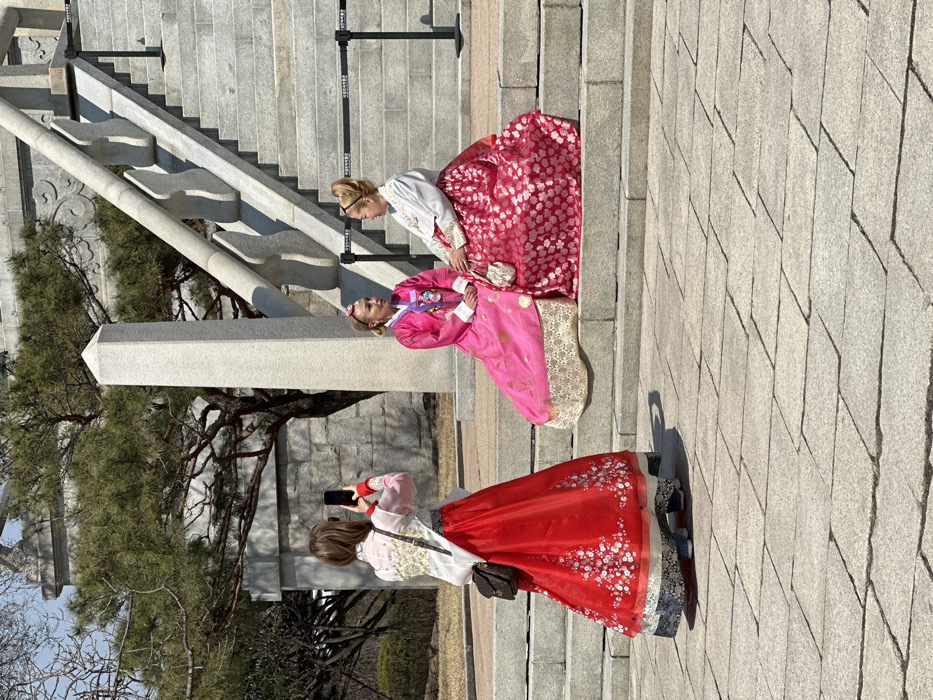 Three female subjects are depicted outdoors at the National Palace Museum of Korea, situated within the Gyeongbokgung Palace grounds in Jongno-gu, Seoul, South Korea. The scene is set against a large, multi-tiered stone staircase with decorative balustrades and a paved stone courtyard.
In the foreground, a female subject with light brown hair, wearing a white embroidered traditional Korean hanbok top over a red floral-patterned hanbok skirt, stands with her back to the camera. She holds a dark smartphone vertically, apparently photographing the two other subjects. A dark brown cross-body bag hangs from her shoulder.
Two other female subjects, both with light hair styled in updos with pink accessories, are seated on the stone steps to the right. The subject on the viewer's left wears a vibrant pink hanbok with gold and white embroidery. The subject on the viewer's right wears a white hanbok top with gold trim and a red floral-patterned hanbok skirt, holding a small, ornate, light-colored handbag. Both seated individuals are positioned to appear as if interacting with each other while being photographed.
The background features stone architectural elements, including a carved stone wall and a large pine tree with green foliage. A tall, light grey stone obelisk-like pillar is visible to the left of the stairs. Black stanchions with retractable barriers, bearing the Korean text "국립고궁박물관" (National Palace Museum of Korea), are present on the steps. The sky is clear and blue, suggesting bright daylight. The ground is paved with rectangular light grey stones.