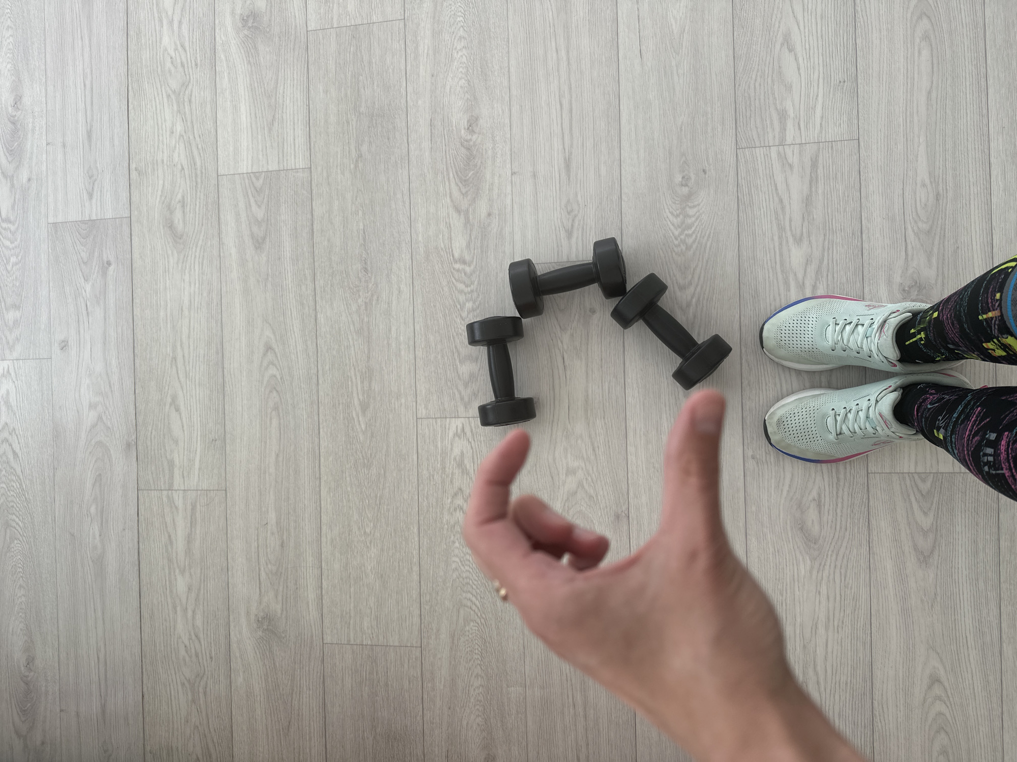 The image depicts a person standing on a light-colored, wood-grain floor, possibly laminate or vinyl flooring. The person is wearing running shoes in a light blue-gray color with colorful accents and black leggings with a colorful print. A hand is partially visible in the foreground, giving a thumbs-up. Three black dumbbells are arranged near the center of the frame. The location appears to be indoors, possibly a home or exercise studio, in the city of Unknown, Unknown.