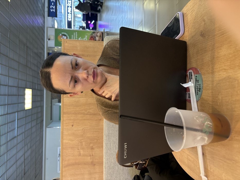 A woman, appearing to be in her 30s-40s with dark hair pulled back, is seated at a light wooden table, facing an open, dark-colored CHUWI laptop. She wears a brown knitted top and gazes at the laptop screen with a serious or concerned expression, her right hand touching her neck. On the table, to the left of the laptop, is a clear plastic cup containing a partially consumed dark liquid and a black straw. Two white paper straws, one unwrapped, lie beside the cup. A circular coaster with Russian text and a QR code is beneath the straws. A smartphone with a dark screen and a light purple case rests to the right of the laptop. The scene is an indoor commercial or public space in Russia, indicated by Russian signage, featuring light wooden partitions, upholstered seating, and a grid ceiling with overhead lighting. In the background, blurry figures are visible near various commercial kiosks, including one labeled "cas-mo-ice" and another with "КАРТОШКА." The specific location within the city of Novogorelovo, Russia, cannot be precisely identified from the image.