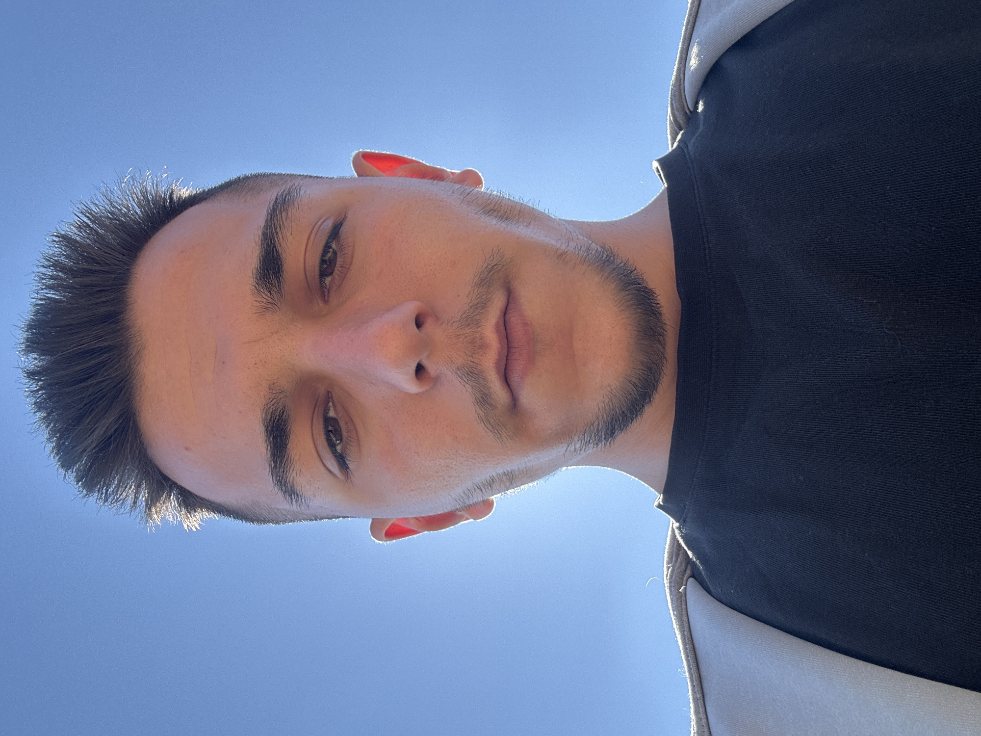 A selfie taken from a low angle. A person is facing the camera, with a short haircut and a goatee. The person is wearing a black t-shirt, and what appears to be a light-colored hooded jacket or sweatshirt. The background is a clear, bright blue sky. The location in the city of Srednia, Bulgaria cannot be ascertained from the image.
