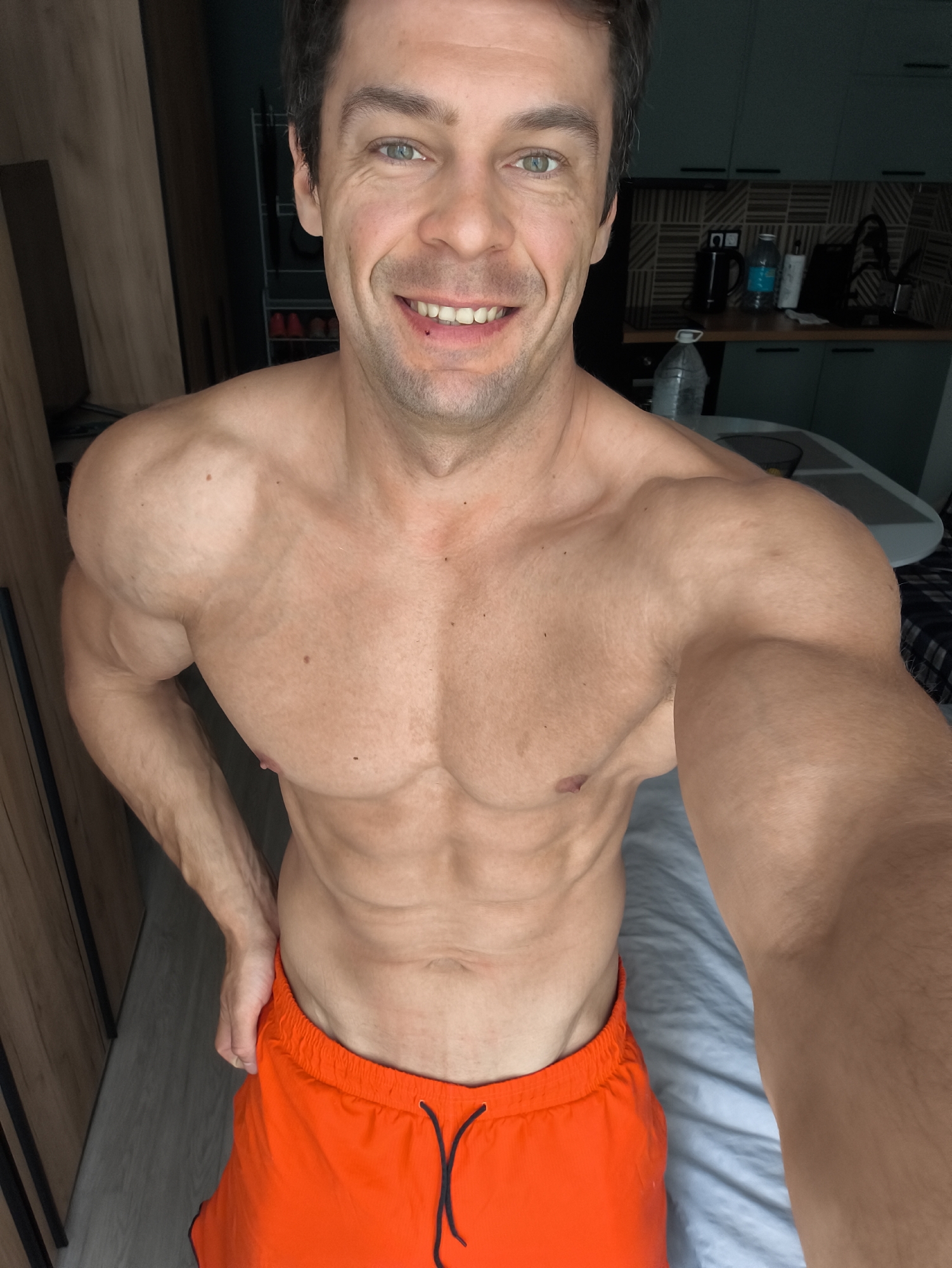 A muscular man with short dark hair and green eyes is taking a selfie indoors. He is shirtless, wearing bright orange shorts, and smiling. The background shows a room with wooden cabinets, a shelf unit, and a kitchen area with blue cabinets and a table with items like bottles and a kettle. The location appears to be a residential interior, but it cannot be specifically determined to be in Moscow, Russia, based on the provided image.