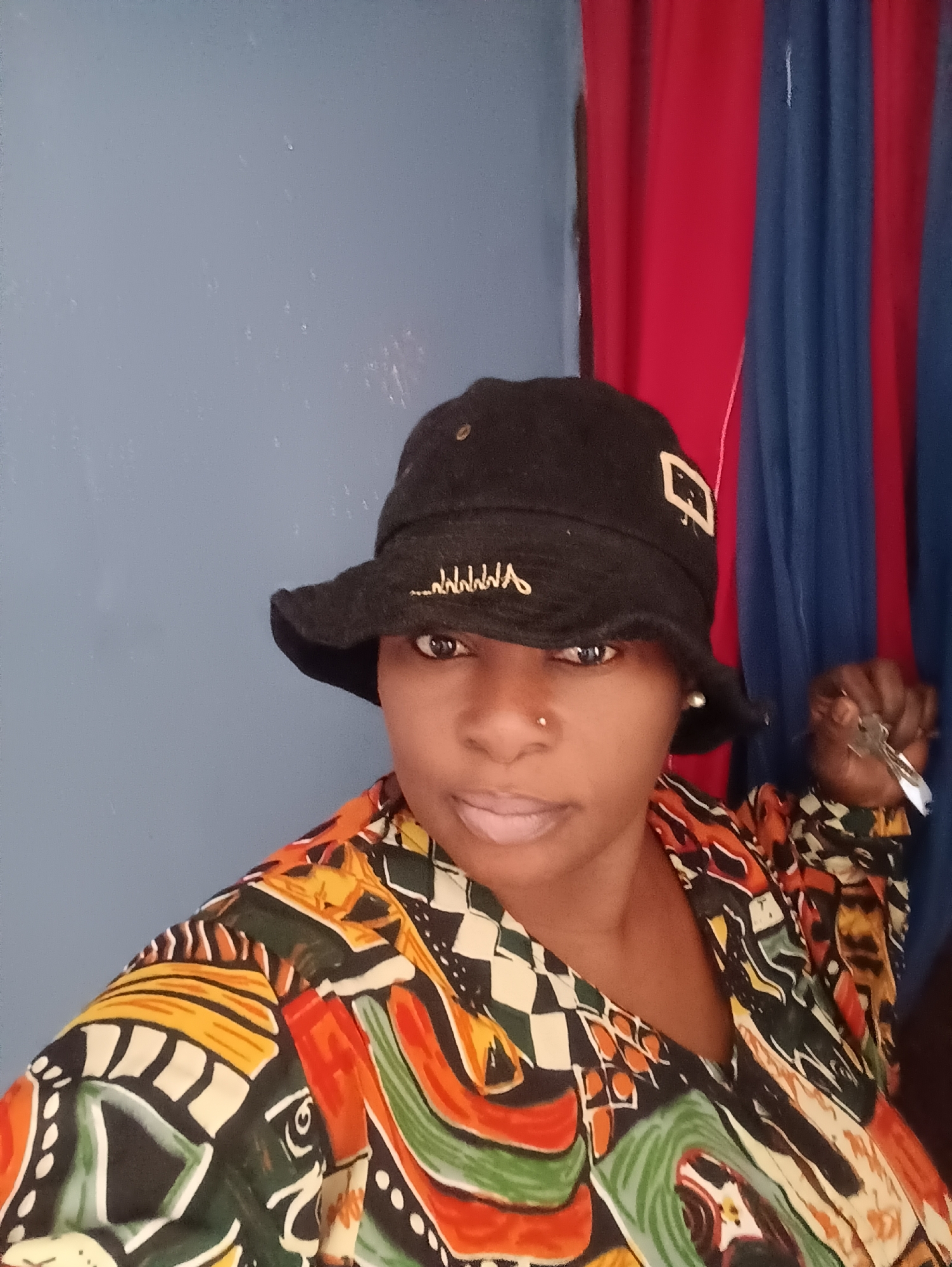 A woman is posing for a selfie indoors. She is wearing a black bucket hat with white embroidery and a colorful, patterned shirt. She has a nose piercing and is holding keys in her right hand. Behind her, there is a blue wall and a section of red and blue curtains. The photograph appears to be taken in Jos, Nigeria.