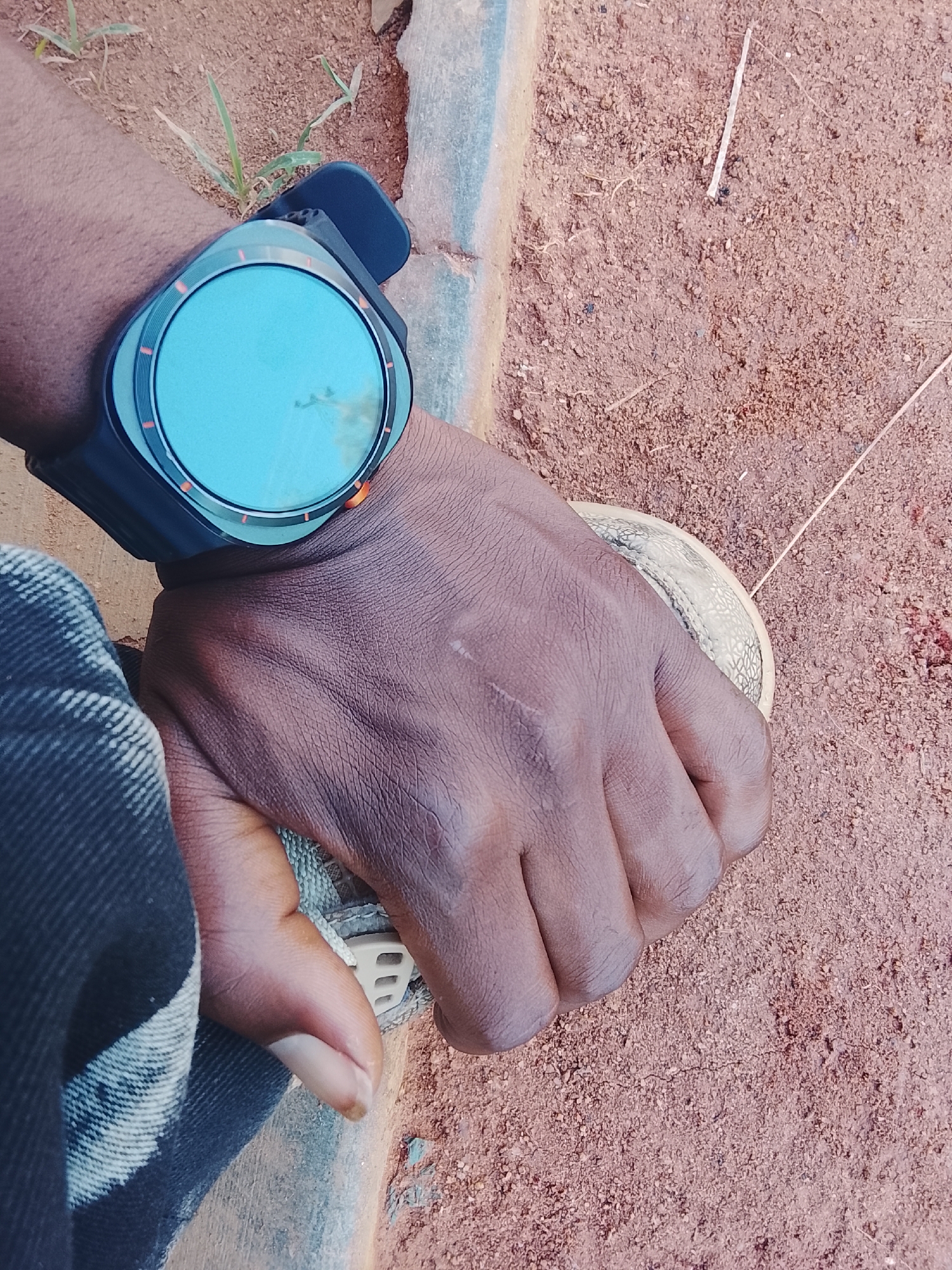 A close-up, over-the-shoulder shot shows a person's left hand and arm, wearing a smartwatch with a blue face and a black band, resting on the ground. The ground is composed of reddish-brown dirt with scattered pebbles and a few blades of dry grass. A portion of what appears to be a concrete or stone border is visible on the left side of the image, with a dark gray, textured fabric, possibly jeans, draped over it. The hand is partially curled around a light-colored, circular object with a textured surface, possibly a small ball or a part of a shoe. The lighting suggests an outdoor environment, likely during the daytime, casting subtle shadows. The image is taken from a high angle, looking down at the hand and wrist.