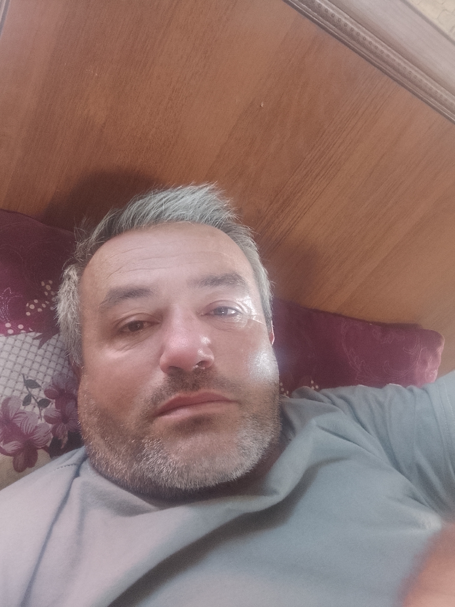 A man with grey hair and a beard is lying down in what appears to be a bed. The background shows a wooden headboard. The man is wearing a grey t-shirt. The pillow he is resting on has a floral pattern. The scene appears to be indoors, likely in a bedroom. No specific location in Sabir Rakhimov, Uzbekistan, is discernible from the image.
