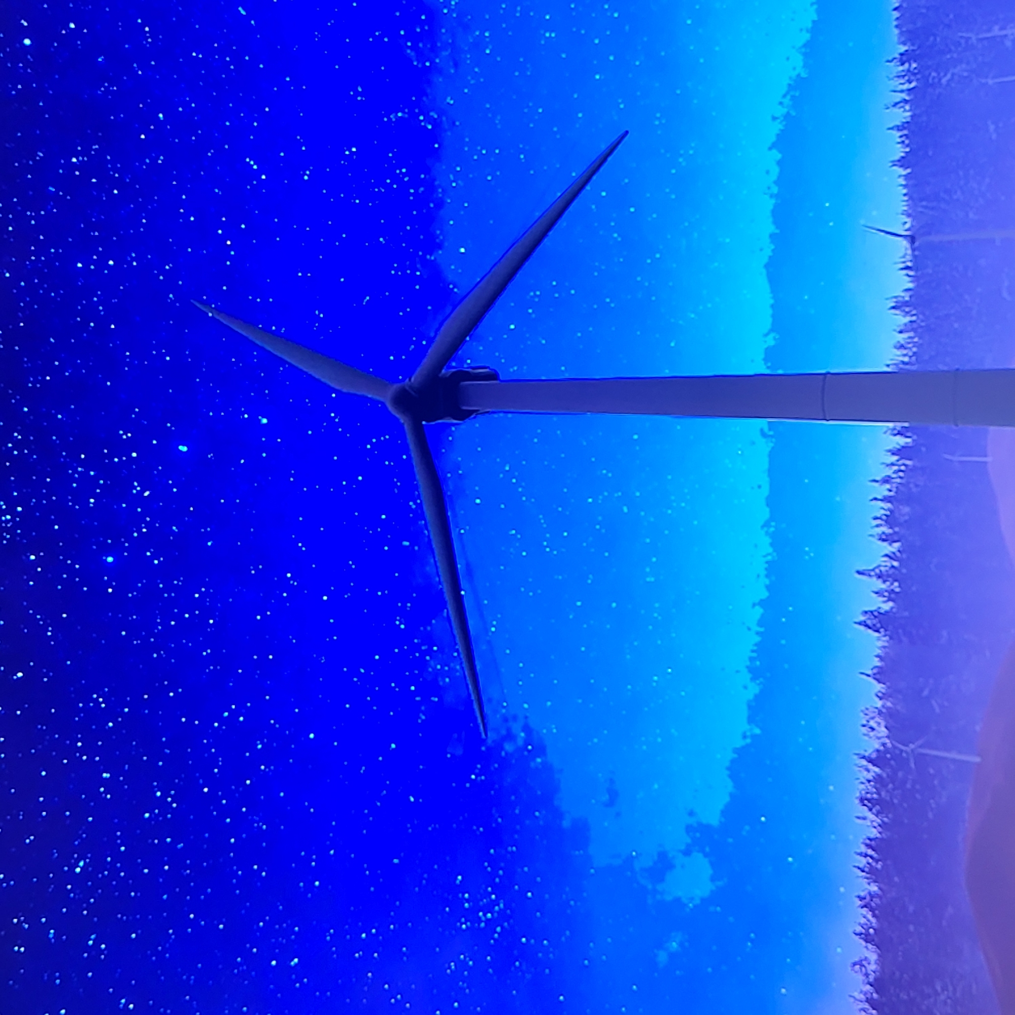 The image shows a wind turbine against a background of blue sky, with a dark blue portion appearing to mimic a starry night effect. A strip of trees is visible on the right side. The location is unknown.