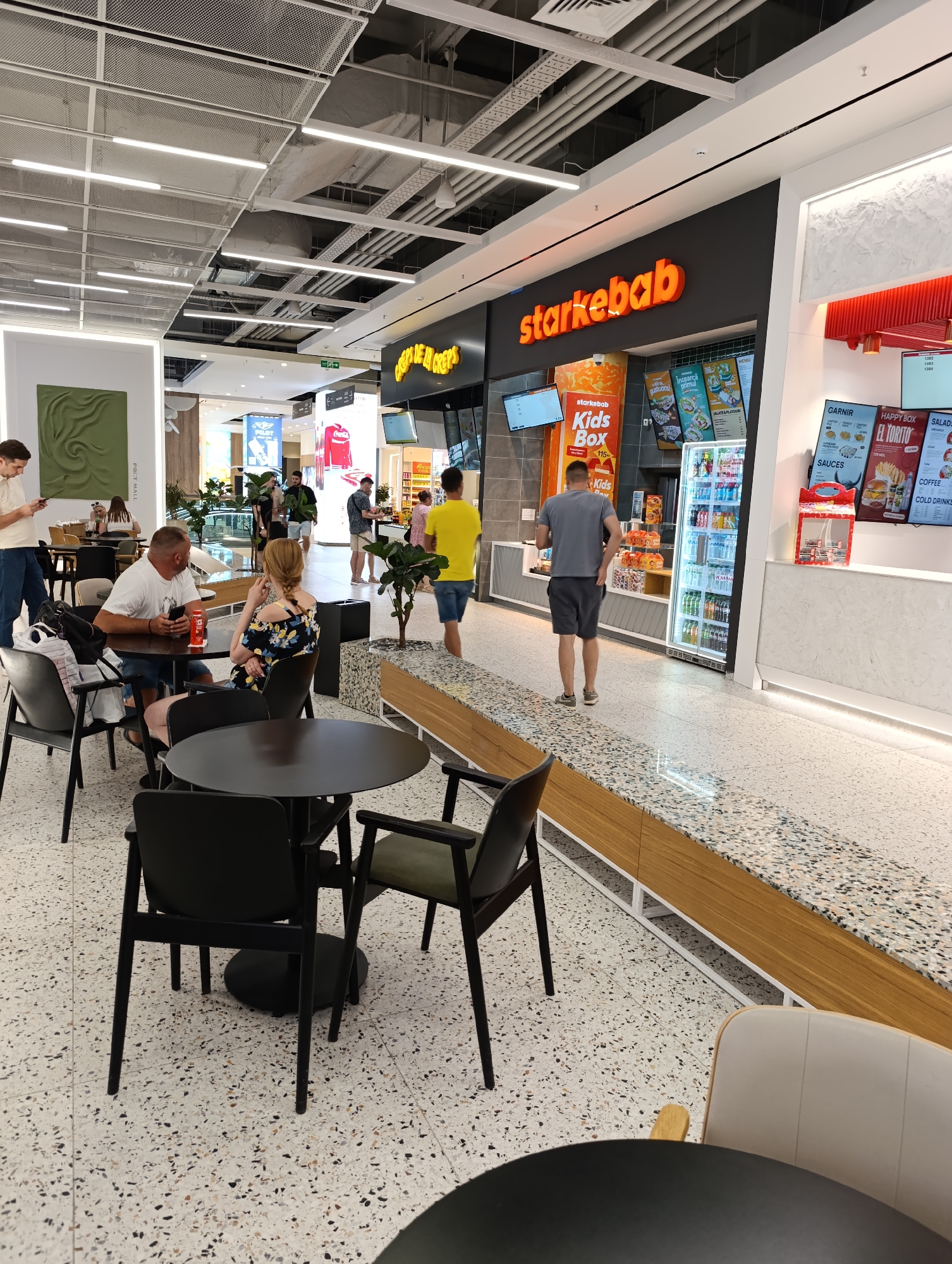A wide-angle, eye-level, outdoor shot captures a contemporary food court, likely within a shopping mall in Chișinău, Moldova. The scene is dominated by a brightly lit fast-food establishment named "starkebab" with an illuminated orange sign. Customers are seen queuing and interacting at the counter, while others are seated at black tables and chairs arranged on a terrazzo-patterned floor. A man in a grey t-shirt and shorts walks away from the counter towards the seating area. A woman in a yellow t-shirt and blue shorts stands near the counter, facing away from the camera. A couple, a man in a white t-shirt and denim shorts and a woman in a floral dress, are seated at a table in the foreground, engaged in conversation. In the background, other patrons are visible, some walking through the mall. The environment is clean and modern, with fluorescent lighting and decorative elements.