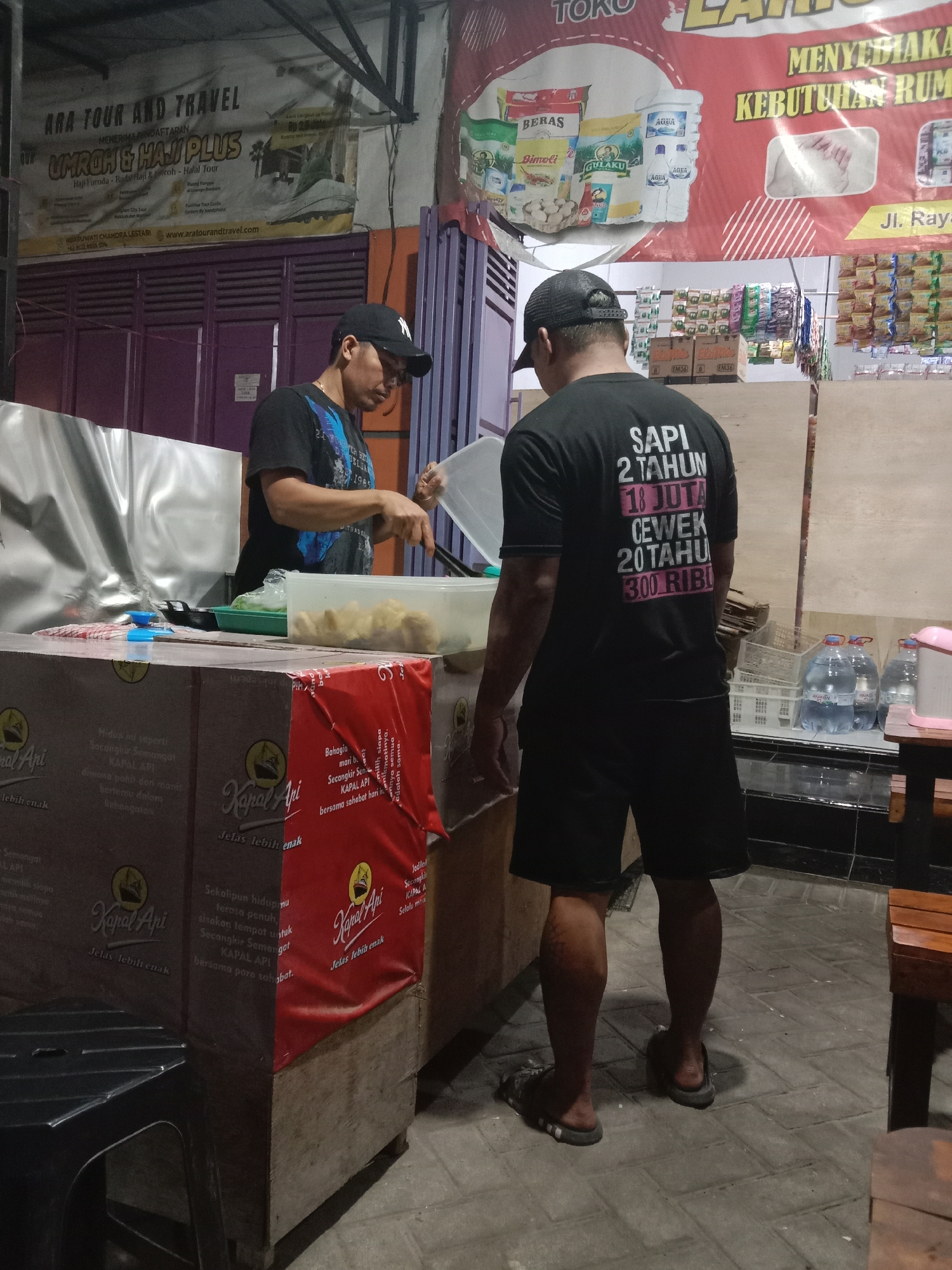In Cangkring, Indonesia, an outdoor street food stall is depicted. A man wearing a black baseball cap and black shorts stands with his back to the camera, facing a food preparation area. He is wearing a black t-shirt with white text on the back that reads "SAPI 2 TAHUN 18 JUTA CEWEK 20 TAHU 300 RIBU". He is holding a clear plastic lid over a white rectangular container filled with food. Another man, wearing a black t-shirt and black baseball cap, is visible from the chest up on the left side of the image, also facing the food preparation area and appears to be engaged in preparing food. The stall is set up with a counter made of wooden boxes covered with red and brown tarps. Various products are displayed behind the men, including packaged snacks, water bottles, and other items. A banner with text in Indonesian advertising "ARA TOUR AND TRAVEL UMROH & HAJJ PLUS" is visible in the background. The ground is paved with light-colored bricks. The lighting suggests it is evening or night.