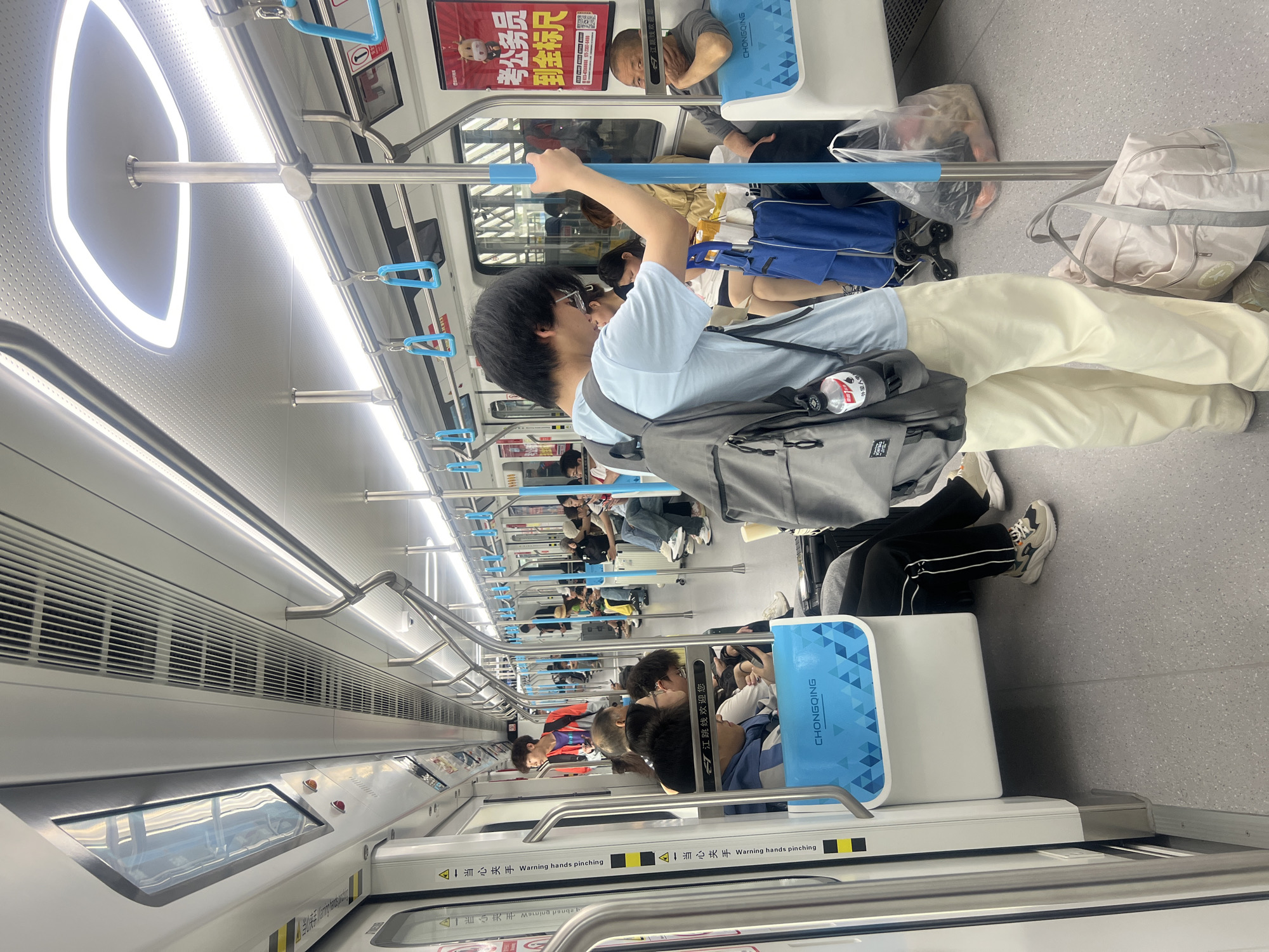 The image shows the interior of a train in Jiangjin, China. There are multiple passengers visible, standing and seated, holding onto handrails. The train car has windows, lighting fixtures, and signage, including warnings about pinching hazards. Seat backs are colored blue with "CHONGQING" printed. Advertisements on the upper portion of the car can be seen above the passengers.