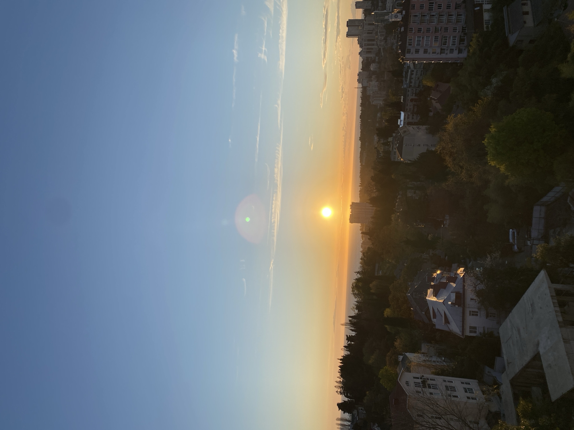 The image depicts a landscape view of what appears to be the city of Sochi, Russia at sunset. The sun is positioned at the horizon line, casting a warm, orange glow across the landscape. The sky transitions from a light blue in the upper left to a yellowish-orange near the horizon. A few wispy clouds are visible in the sky.
Below the horizon, the city is nestled amidst trees and buildings. In the lower right corner, a concrete structure is visible, suggesting the presence of a building under construction or renovation. There are various structures, including residential buildings, visible amongst the trees.
There are no people, actions or interactions visible in the scene.