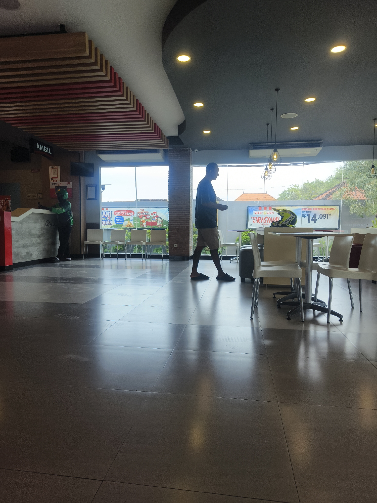 The image depicts the interior of a KFC restaurant in Denpasar, Indonesia. A man in a dark t-shirt and light-colored shorts is walking across the tiled floor, holding a white plate. To his left, near a counter with a red accent, a person in a green uniform, possibly a delivery driver, is interacting with a customer. Behind the counter, there are signs and a television screen. Through a large window, a view of the outdoors is visible, showing trees and buildings. The restaurant features modern lighting fixtures on the ceiling and tables and chairs are arranged for dining. A promotional banner with a motorcycle helmet and text in Indonesian is displayed near the window. The floor is composed of large, dark tiles that reflect the ambient light.