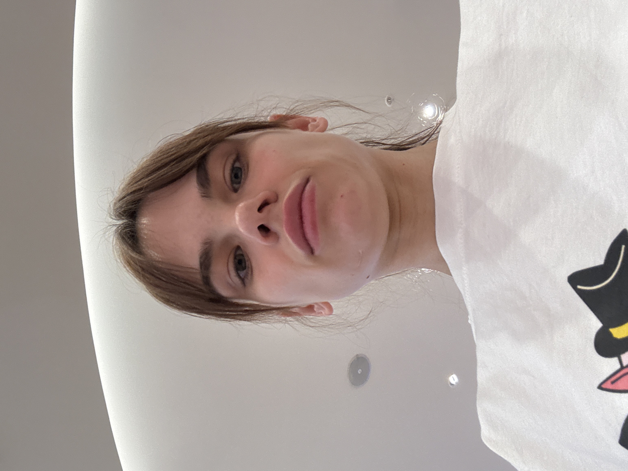 A photograph taken from a low angle shows a fair-skinned woman with brown hair and bangs looking directly at the camera. She is wearing a white T-shirt featuring a cartoon character with a top hat and bowtie. The background appears to be a white ceiling with recessed lighting. The location is Olginskaya, Russia.